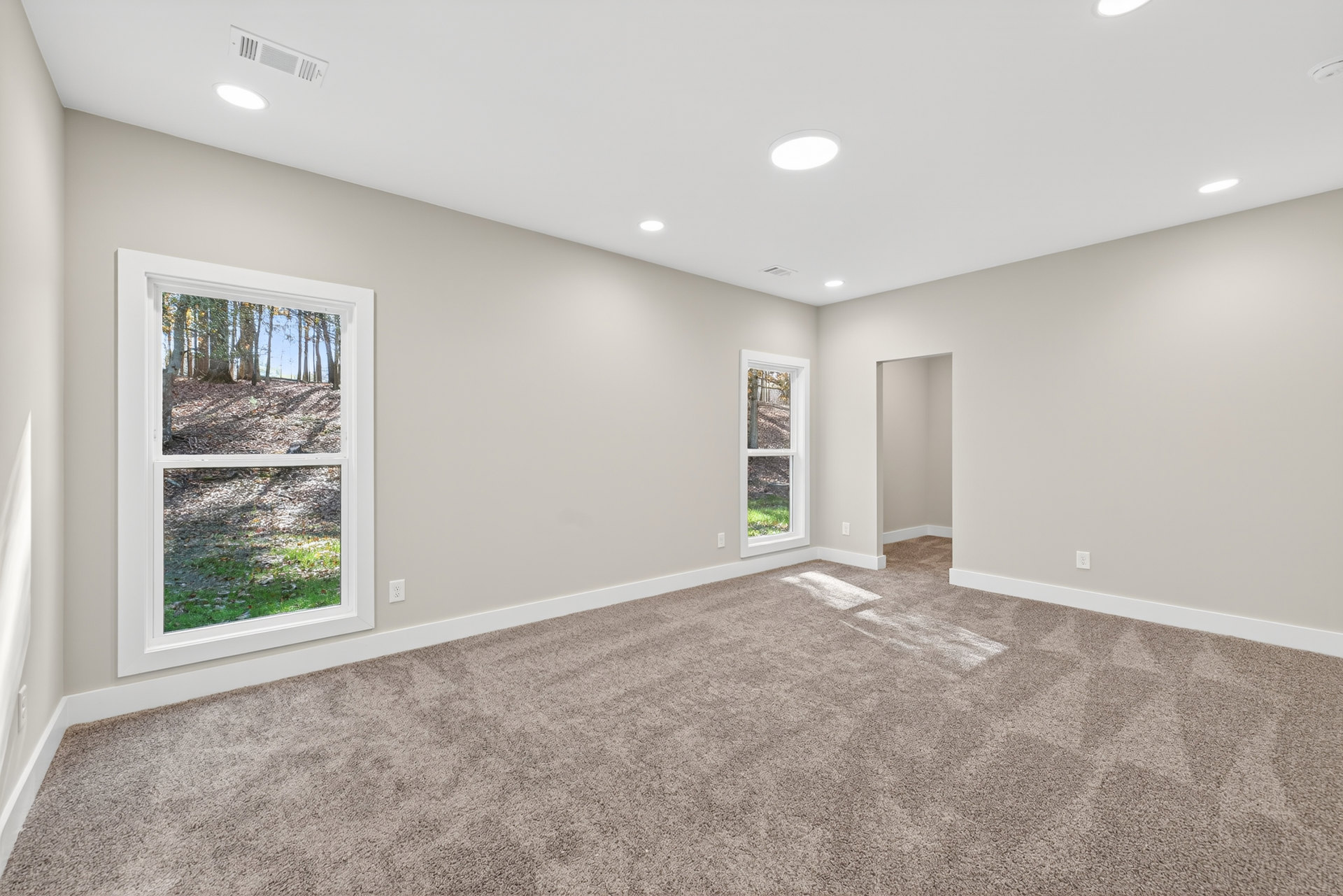 Carpeted room with large windows overlooking forest, white ceiling with recessed lights, neutral walls, and a wooden door