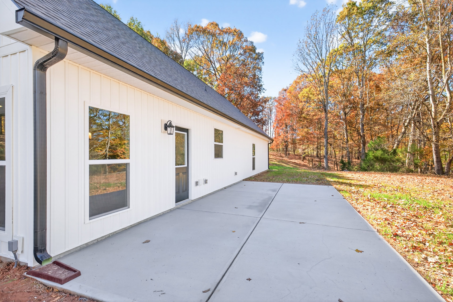 White siding home with black-framed windows, concrete driveway, and mature trees in the background