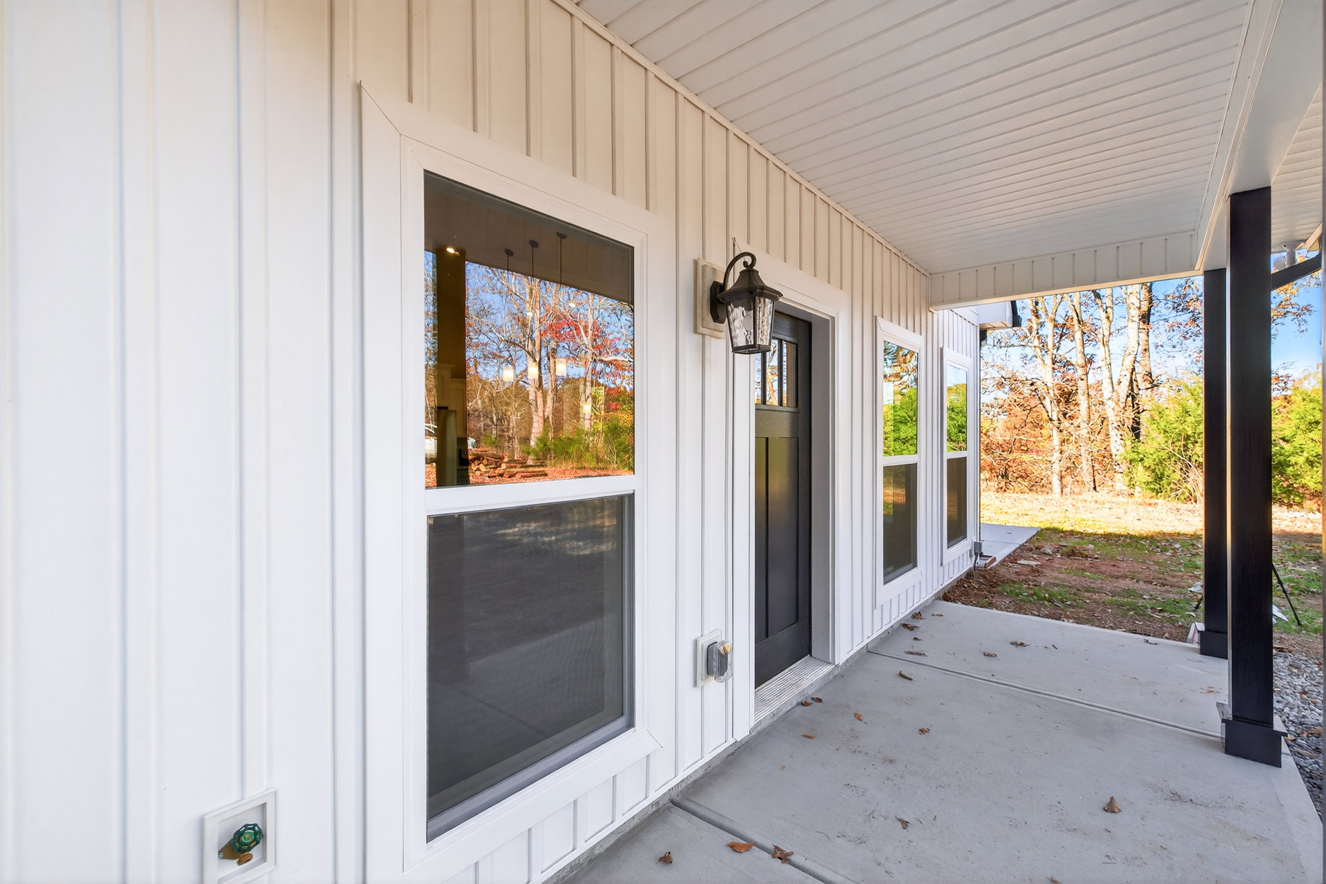 White siding house with black door and windows, covered porch with light fixture, screened window, fallen leaves near entry, trees reflected in glass