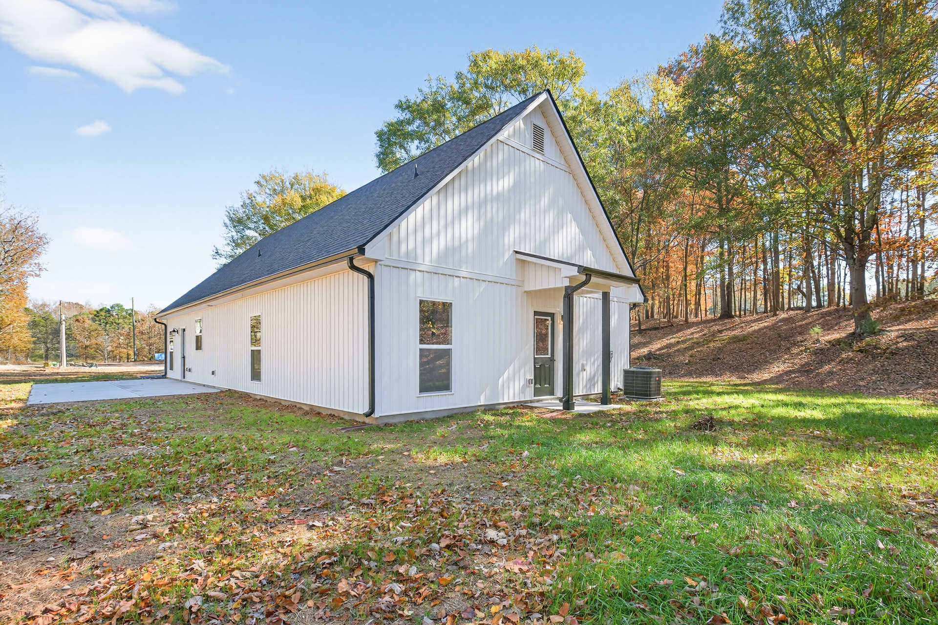 White farmhouse with black roof, large windows, yellow flower in window, black heat pump beside exterior wall, green lawn with scattered leaves, mature trees in background under