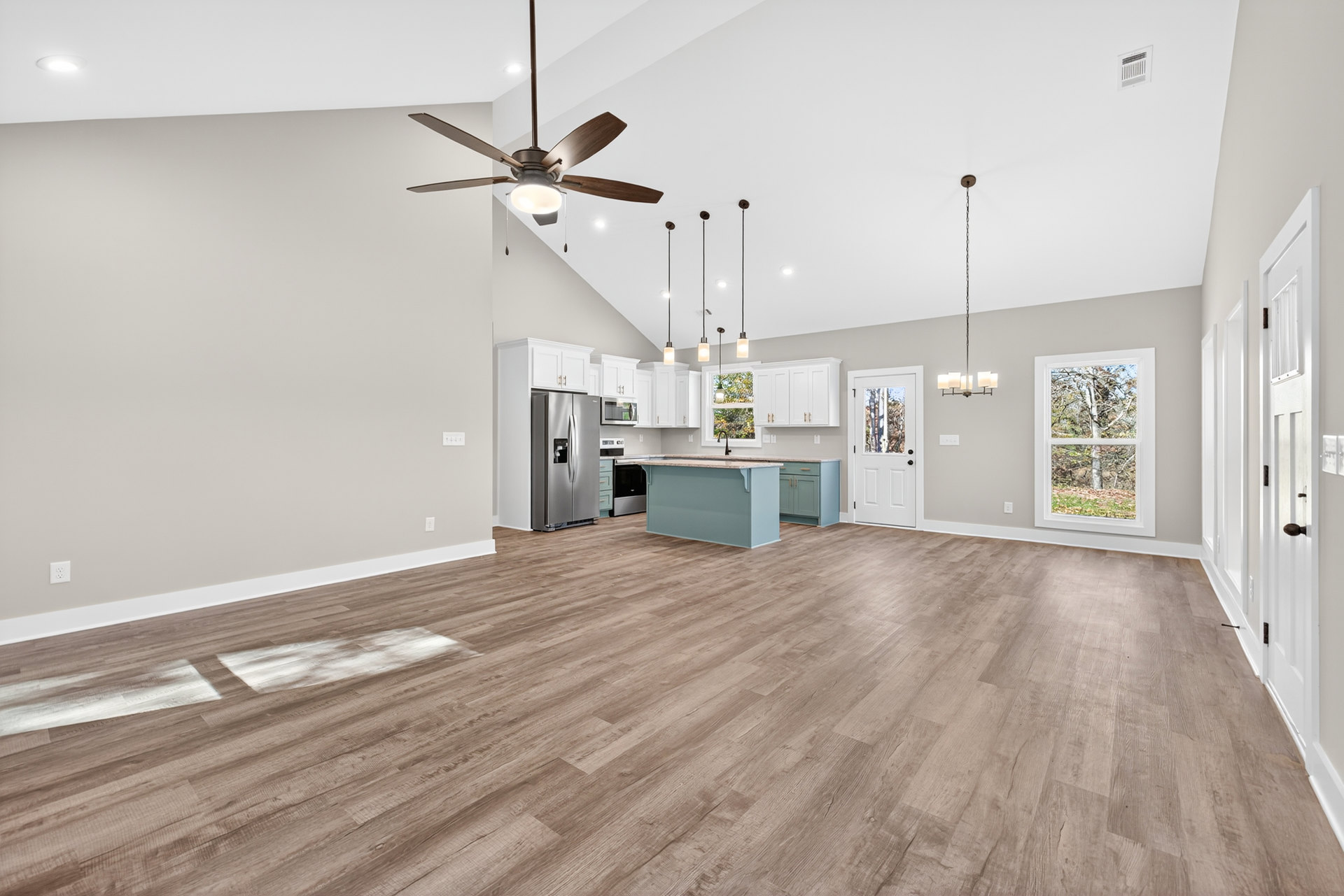 Spacious open floor plan featuring a kitchen with wood flooring, stainless steel refrigerator, ceiling fan with light, large window overlooking trees, and a white door with glass