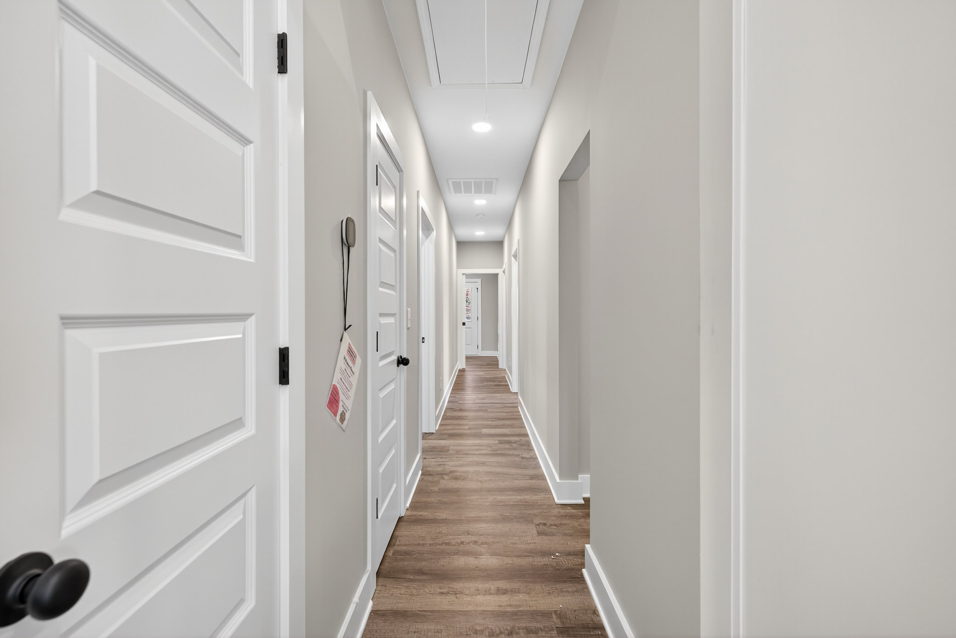 Long hallway with white walls and doors, light wood flooring, recessed ceiling lights, and silver door hardware