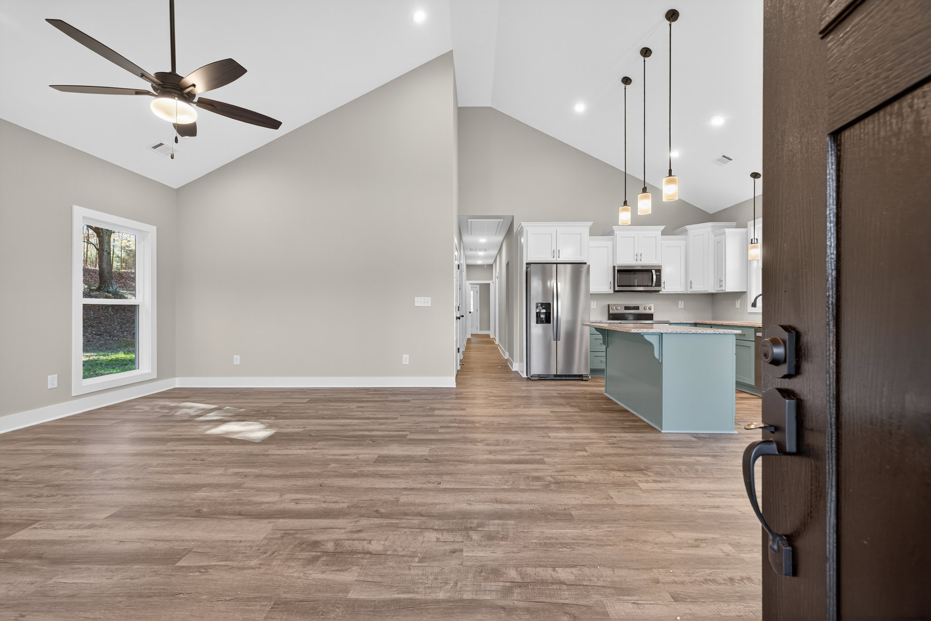 Wide open door leading to kitchen with ceiling fan, stainless refrigerator, window framing tree trunk, and central island with stone countertop