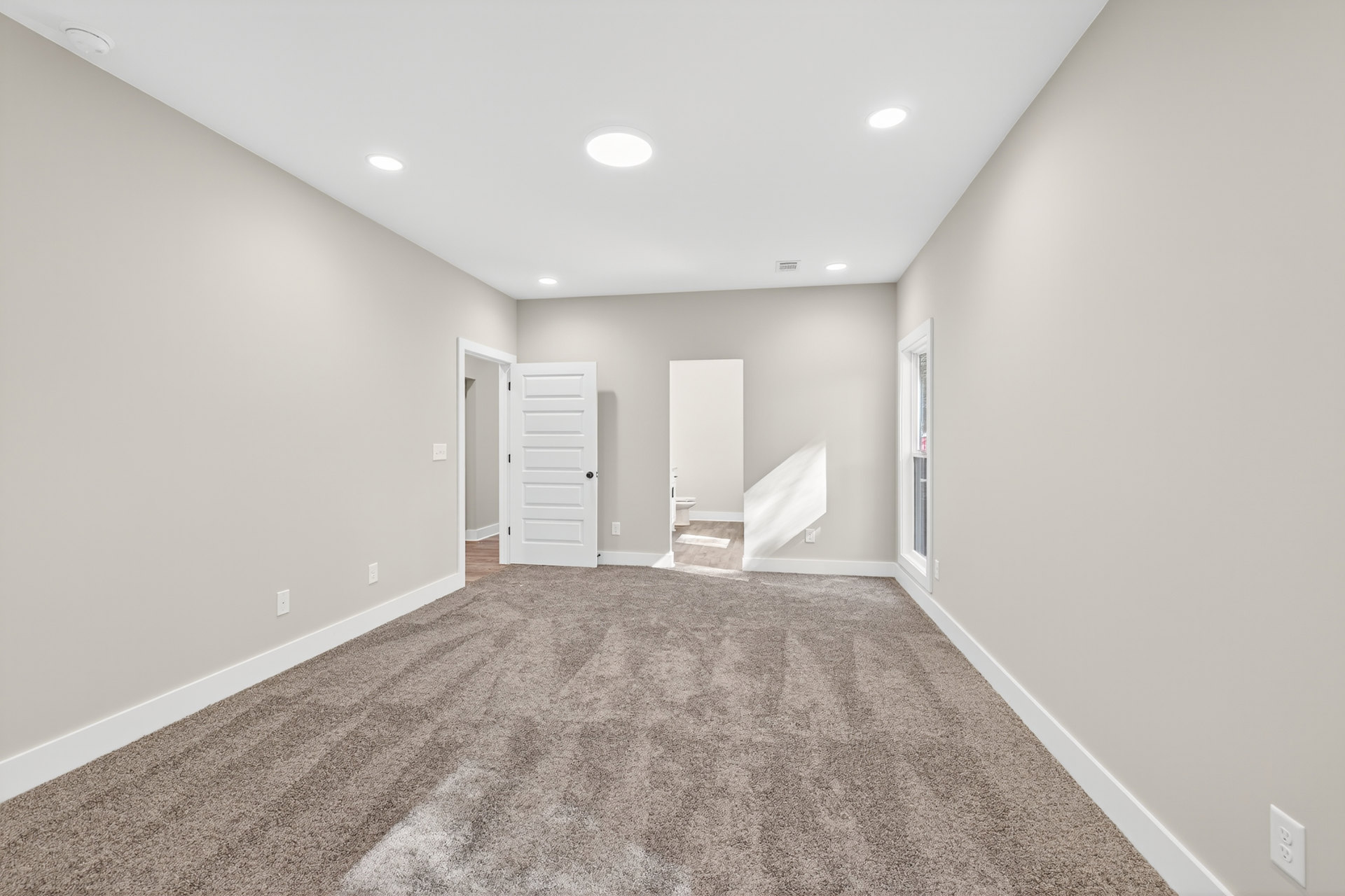 White carpeted room with a white door featuring black knobs, white walls, ceiling light, electrical outlet, and light switch.