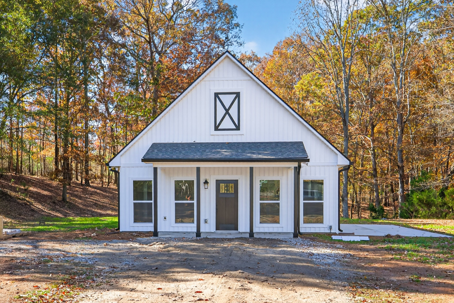 White siding home with black roof, brown front door, large windows, surrounded by trees and greenery under a blue sky