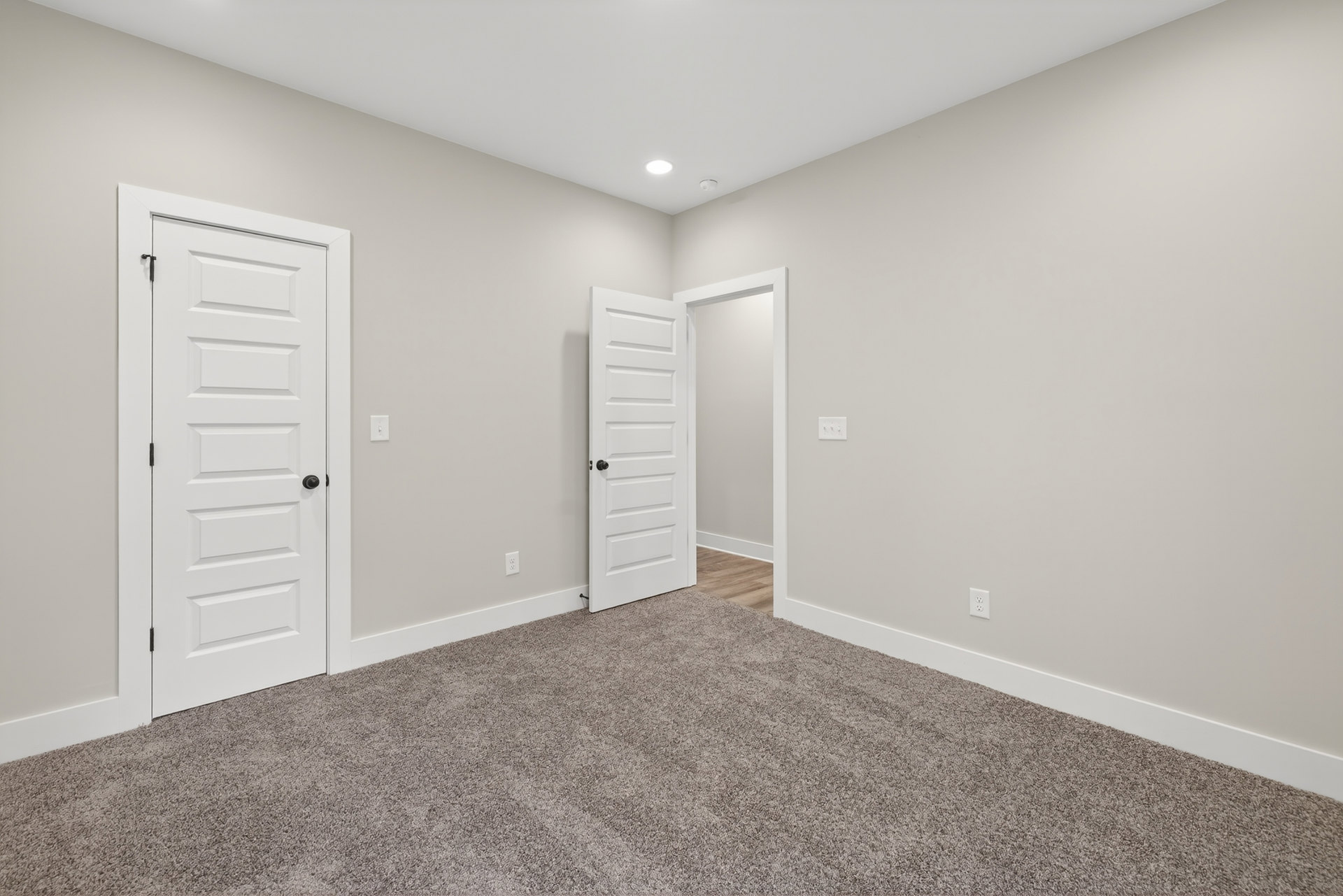White carpeted room featuring white paneled doors with black handles, smooth plaster walls, and a white ceiling with recessed lighting.