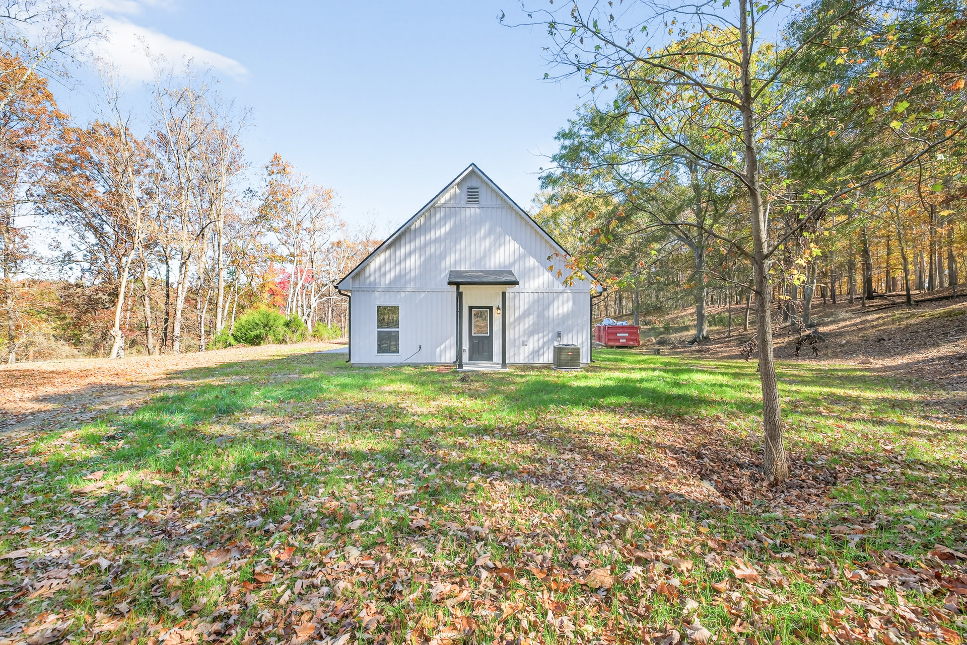 White farmhouse with black door, autumn leaves scattered on lawn, mature trees in background, red dumpster and large metal tank near side of house.