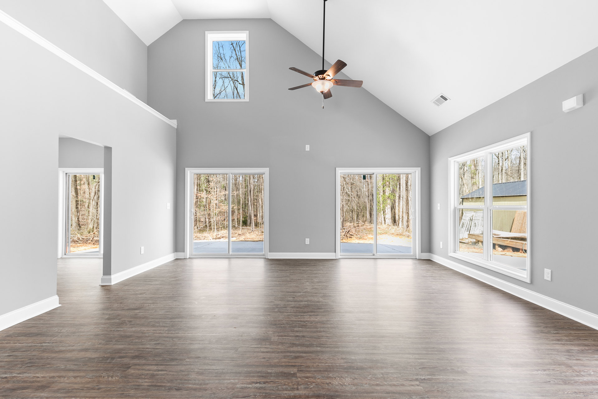 Spacious living area with wood flooring, ceiling fan with light, multiple windows, and sliding glass door overlooking trees