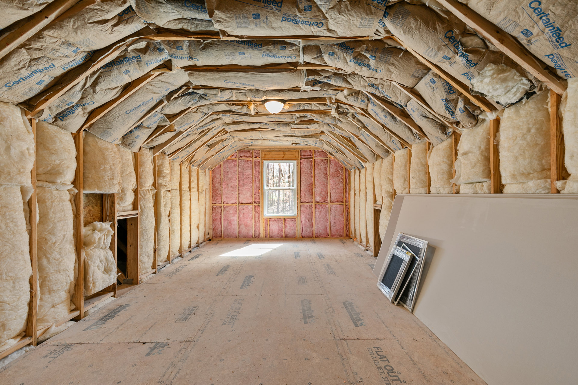 Room with unfinished wood wall, pink insulation, white-framed pictures, sacks stacked along the floor, window showing trees outside, white board mounted on wall.
