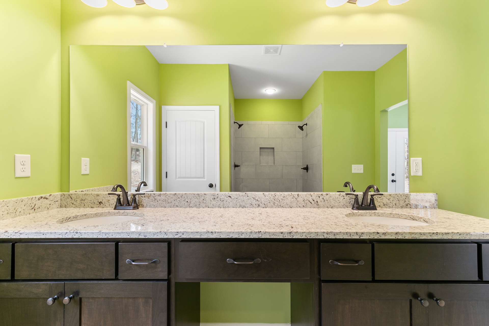 Bathroom with green painted wall, double white sinks set in a stone countertop, wood cabinetry below, large mirror above sinks, white door, window with view of trees, electrical