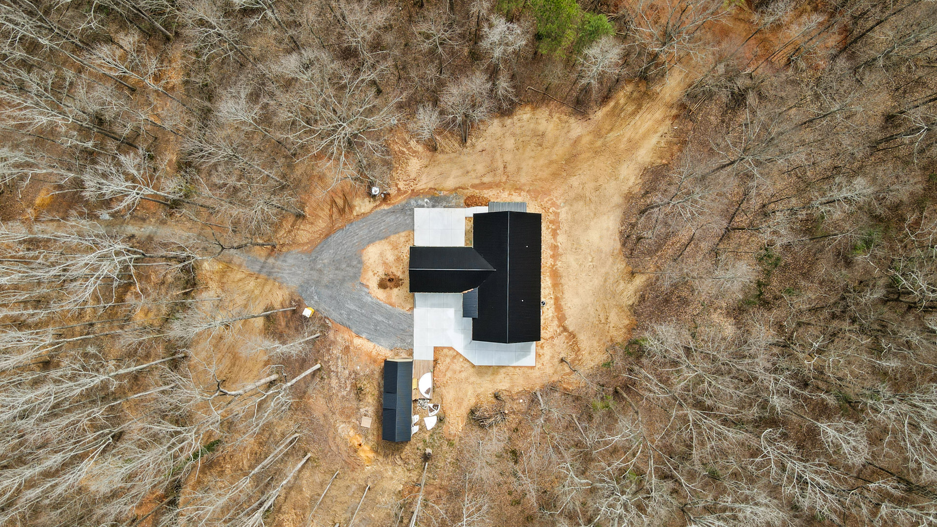 Modern house with black triangular roof, surrounded by mature trees, driveway leading to entrance, tree trunks visible on dirt and grass field