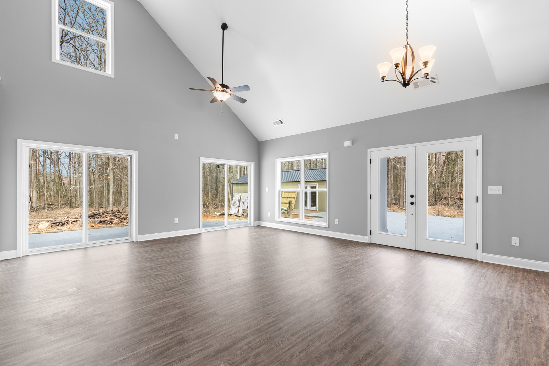 Spacious room with wood flooring, ceiling fan, chandelier, large windows showing tree branches and a road, double glass-paneled doors, plaster walls