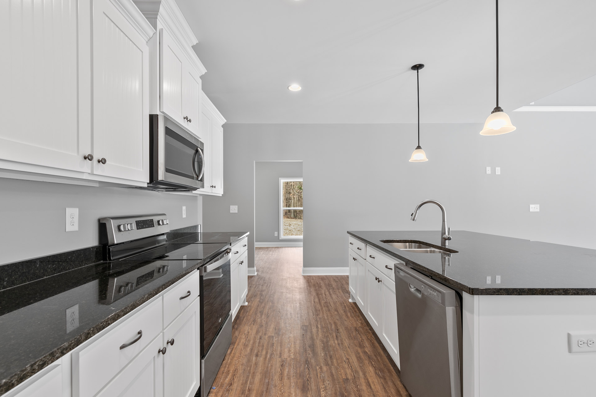 White kitchen cabinets with black countertops, stainless steel microwave, stove, and sink; large window reveals leafy trees outside.