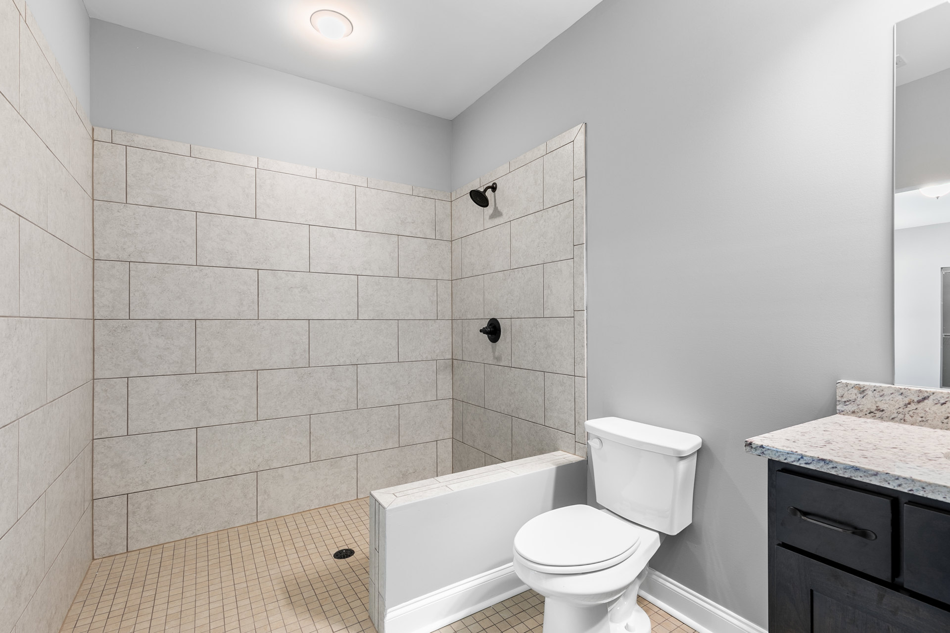 Modern bathroom featuring a white toilet with chrome handle, tiled shower wall with mounted shower head, light fixture above, and vanity with drawer and countertop.