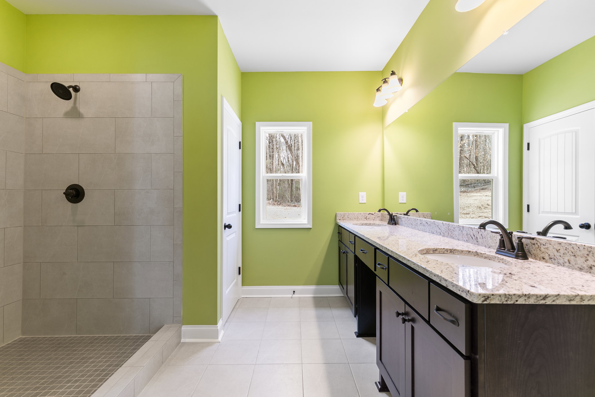 Bathroom with green painted walls, double sink vanity with white countertop and cabinetry, window overlooking trees, round shower head, white door with black hinges, three-light
