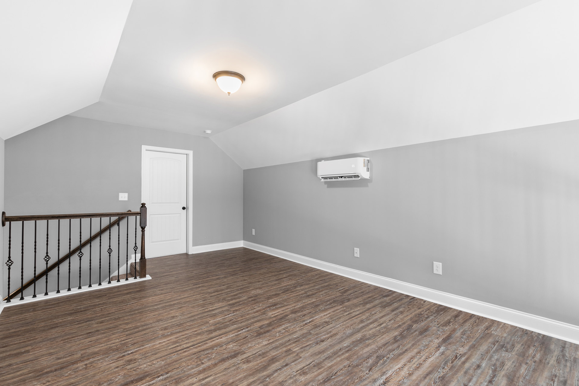 Wood floor with white baseboards, white door featuring black knobs, staircase with wood handrail, ceiling-mounted light fixture, wall-mounted white air conditioner