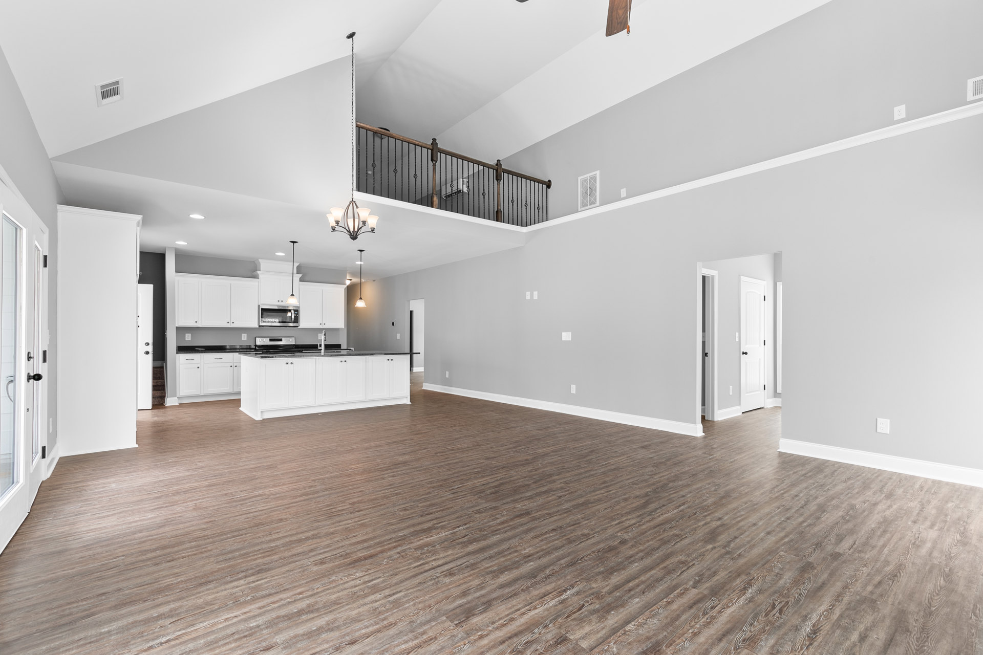 Open-concept kitchen with white cabinets and sink, wood flooring, white wall vent, close-up of metal railing, white rectangular object with black holes on wall