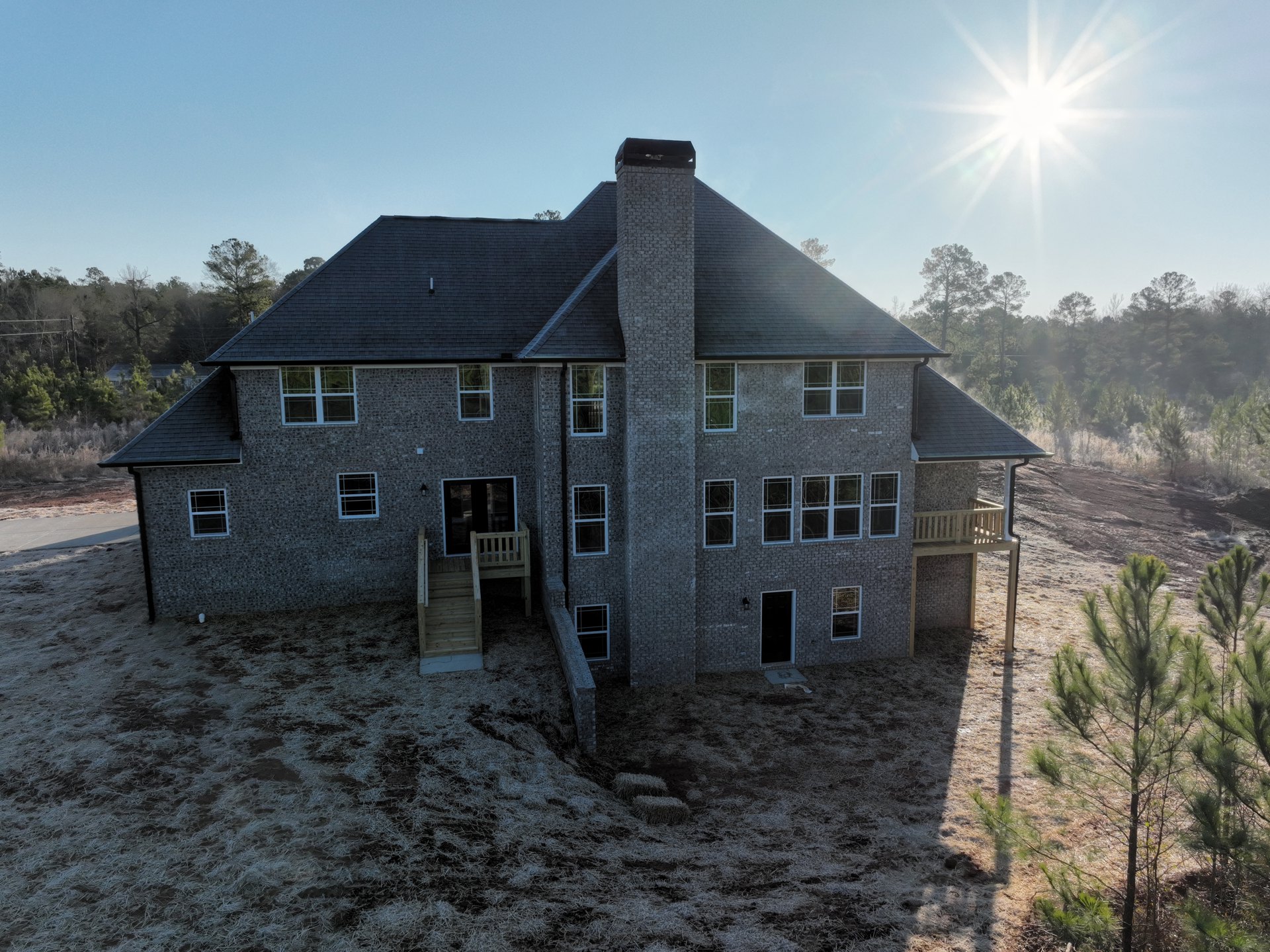 Large brick house with prominent chimney, white-framed windows, surrounded by green grass and mature trees under bright blue sky