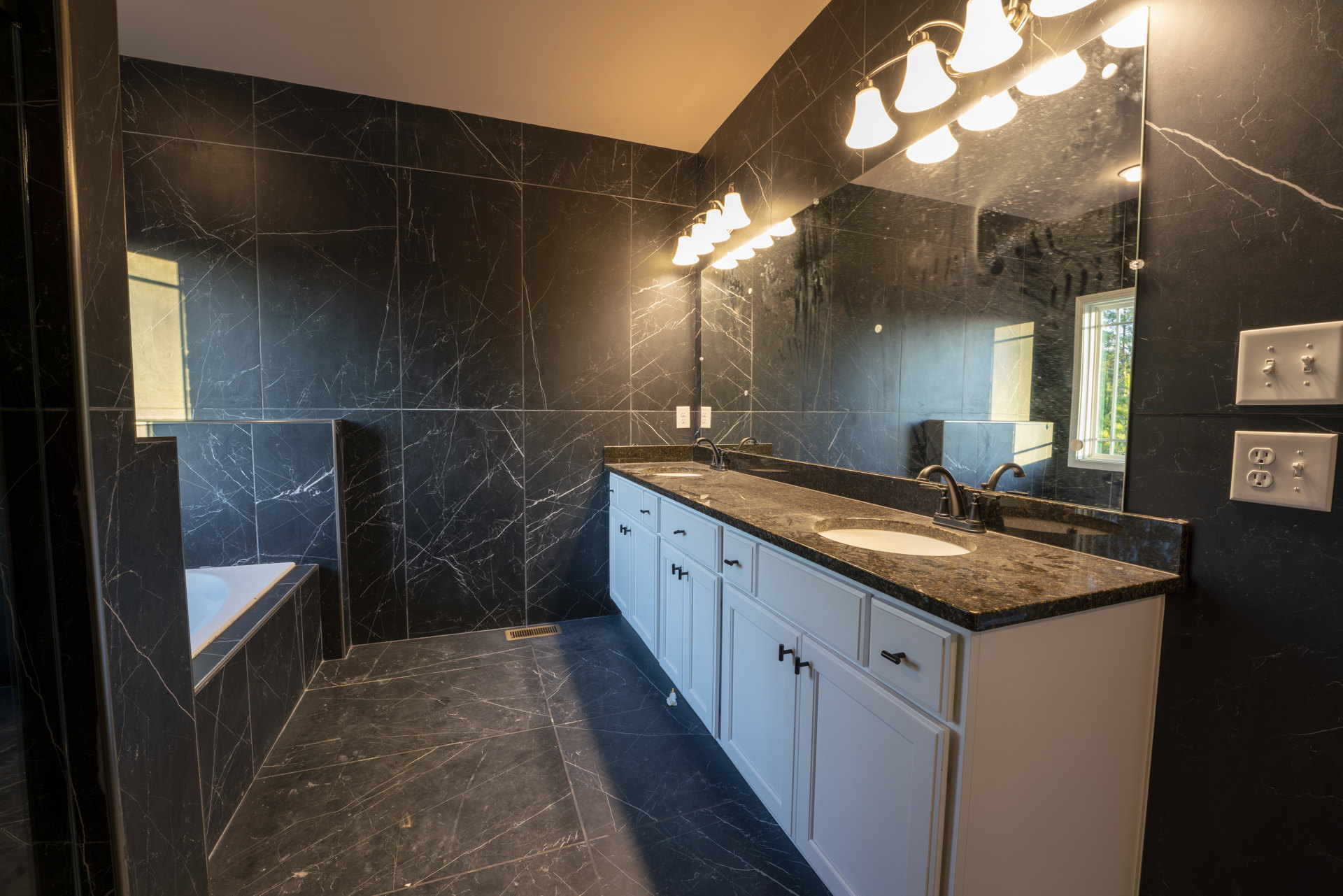 Bathroom featuring marble walls, expansive mirror above marble countertop, modern light fixture, white wall outlet and dual light switch, window revealing trees outside