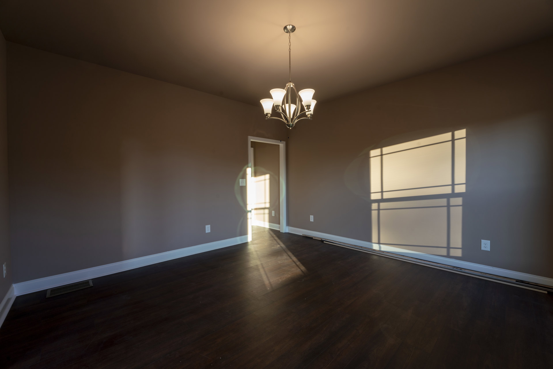 Chandelier with five white lamps hanging from a white ceiling, wood plank flooring, open door, sunlight casting window shadow on pale plaster wall