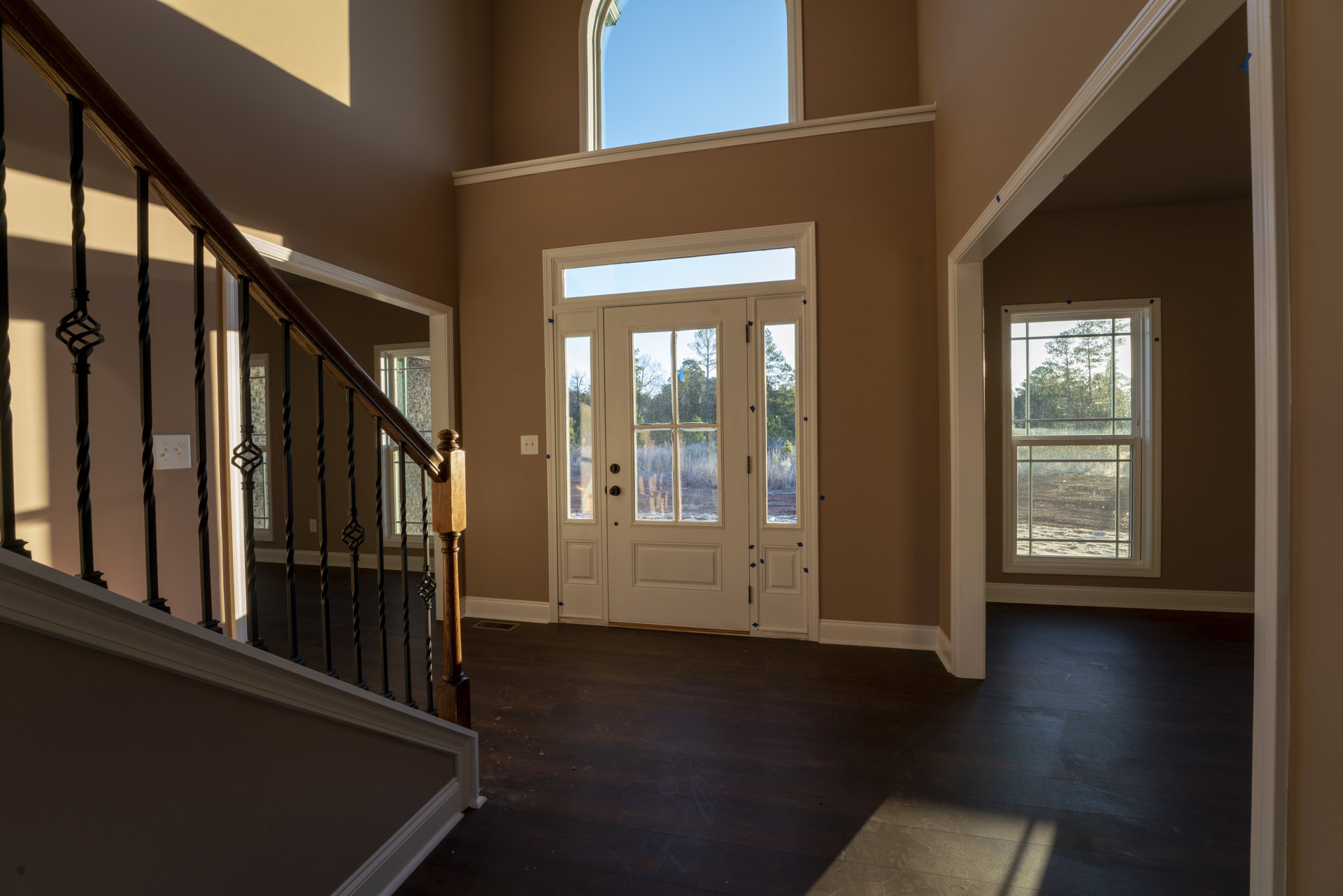 Hallway with hardwood flooring, white staircase with wooden handrail, white double doors with glass panes, window showing trees and blue sky, light switch on wall