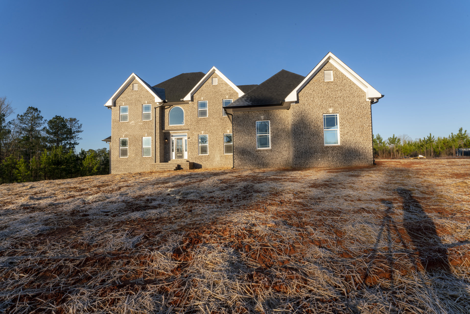Brick house with white-framed windows, gabled roof, expansive grassy yard, mature trees, and clear blue sky