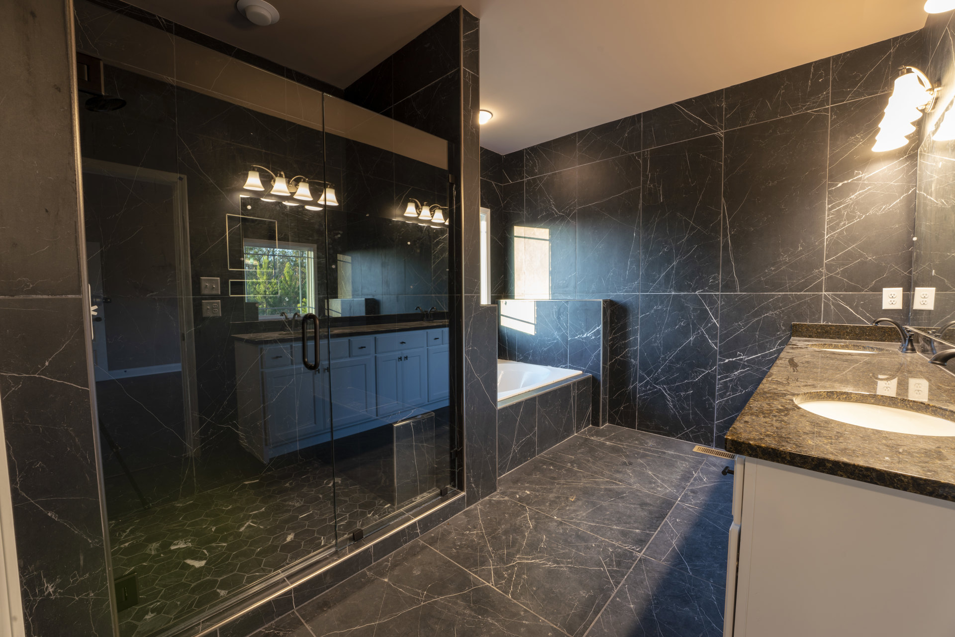 Bathroom with glass shower door, white tile walls, modern sink and faucet, wall-mounted light fixture, large mirror, window overlooking trees