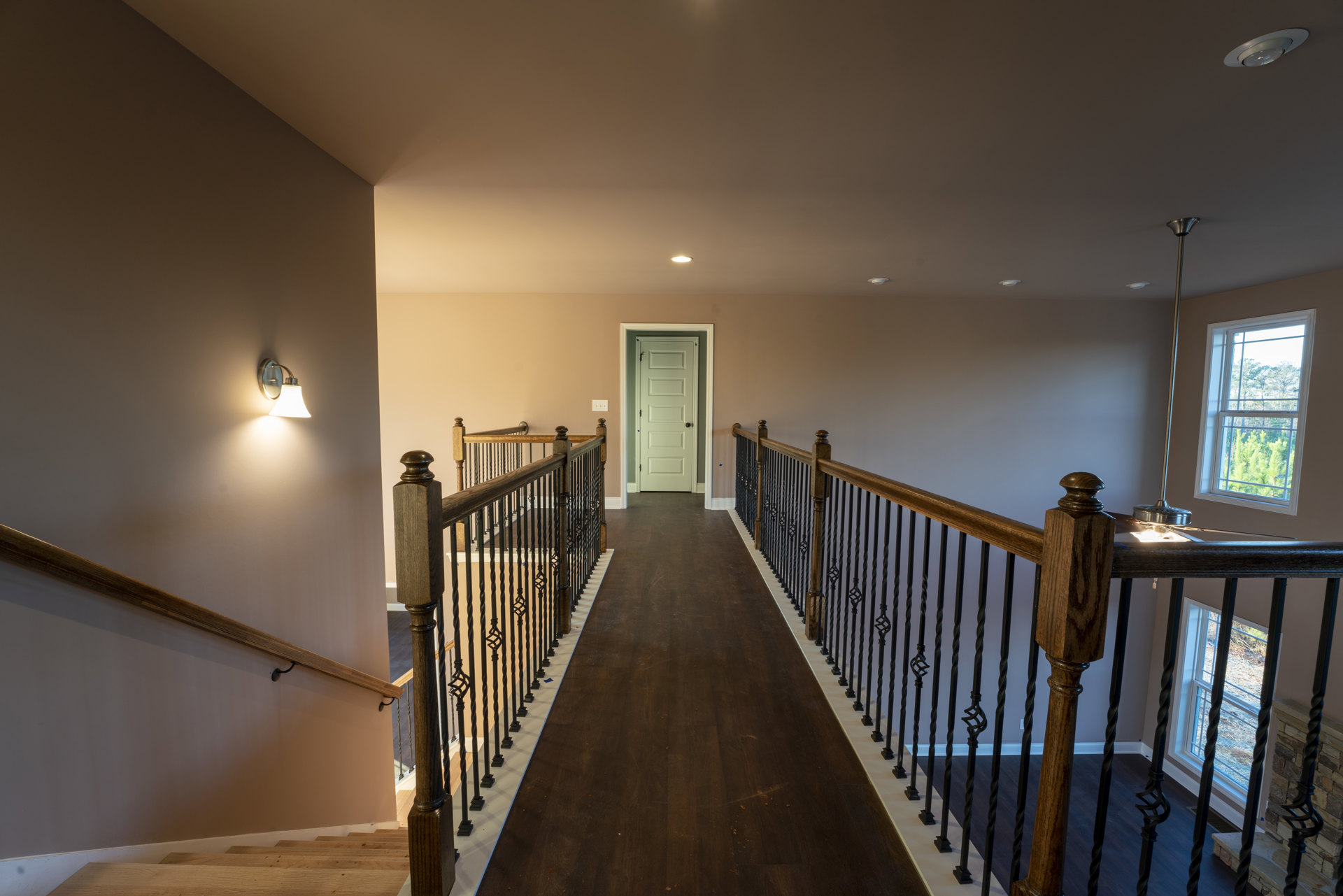 Hallway with warm wood flooring, white door with black knob, metal railings along stairs, wall-mounted light fixture, window revealing trees outside