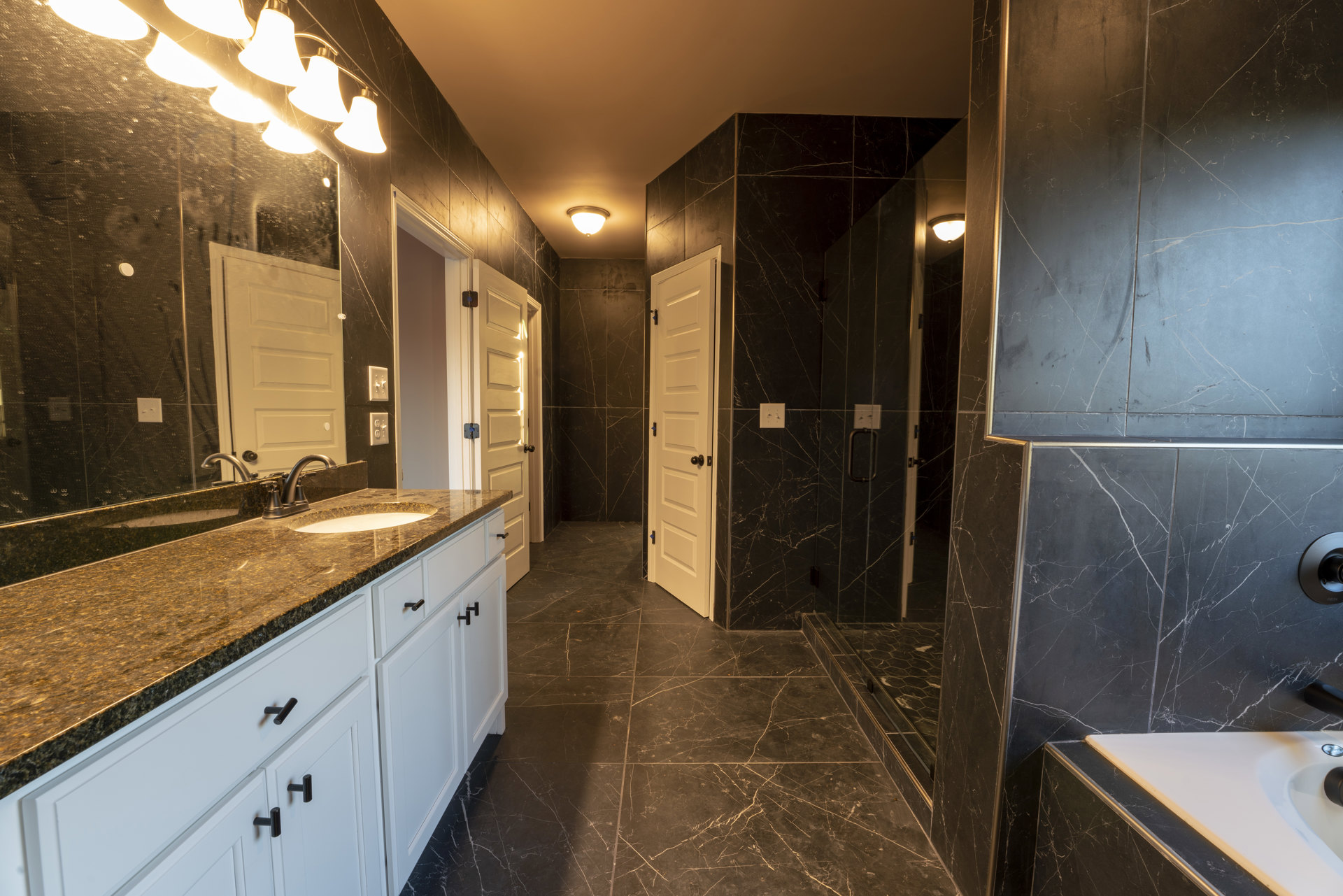 Bathroom with marble wall tiles, white sink set in a bathroom counter with cabinets, corner of a bathtub, white door with black knob, ceiling lights.