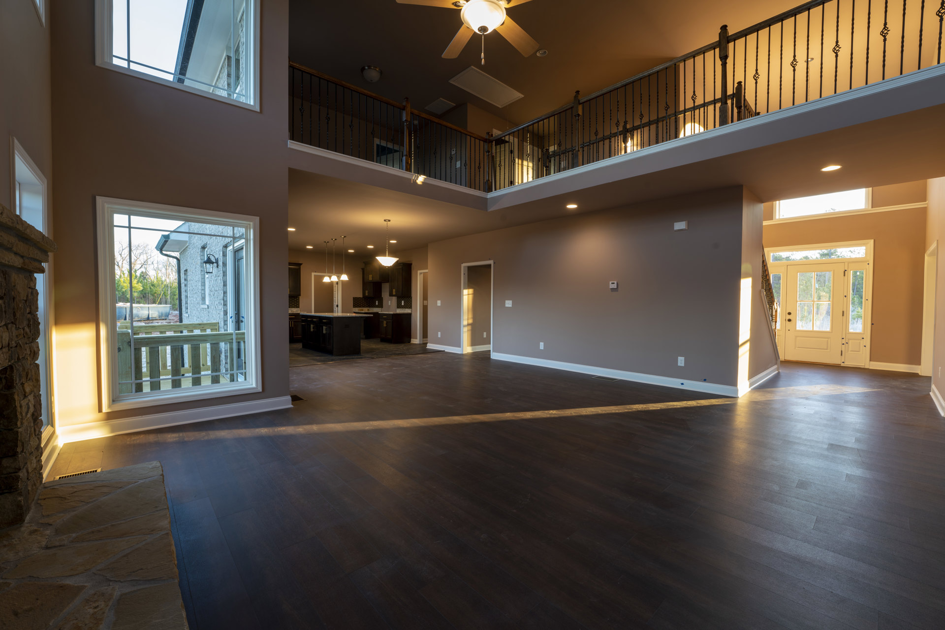 Spacious living area with dark wood flooring, white trim, ceiling fan, large glass doors opening to balcony, and adjacent kitchen with wooden railing
