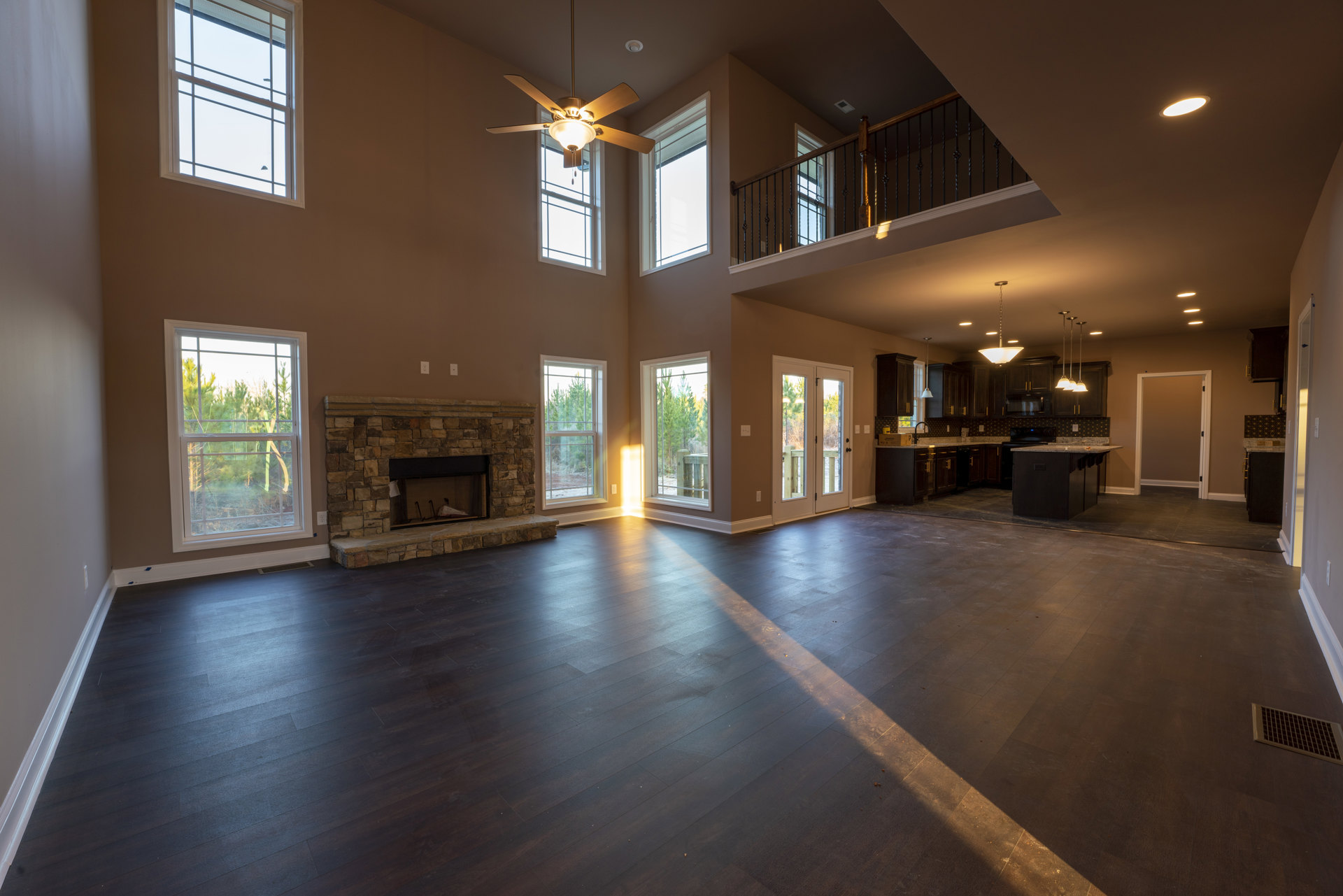 Spacious living room featuring a brick fireplace, ceiling fan with light fixture, hardwood flooring, and open view into a modern kitchen; large window with white frame overlooks