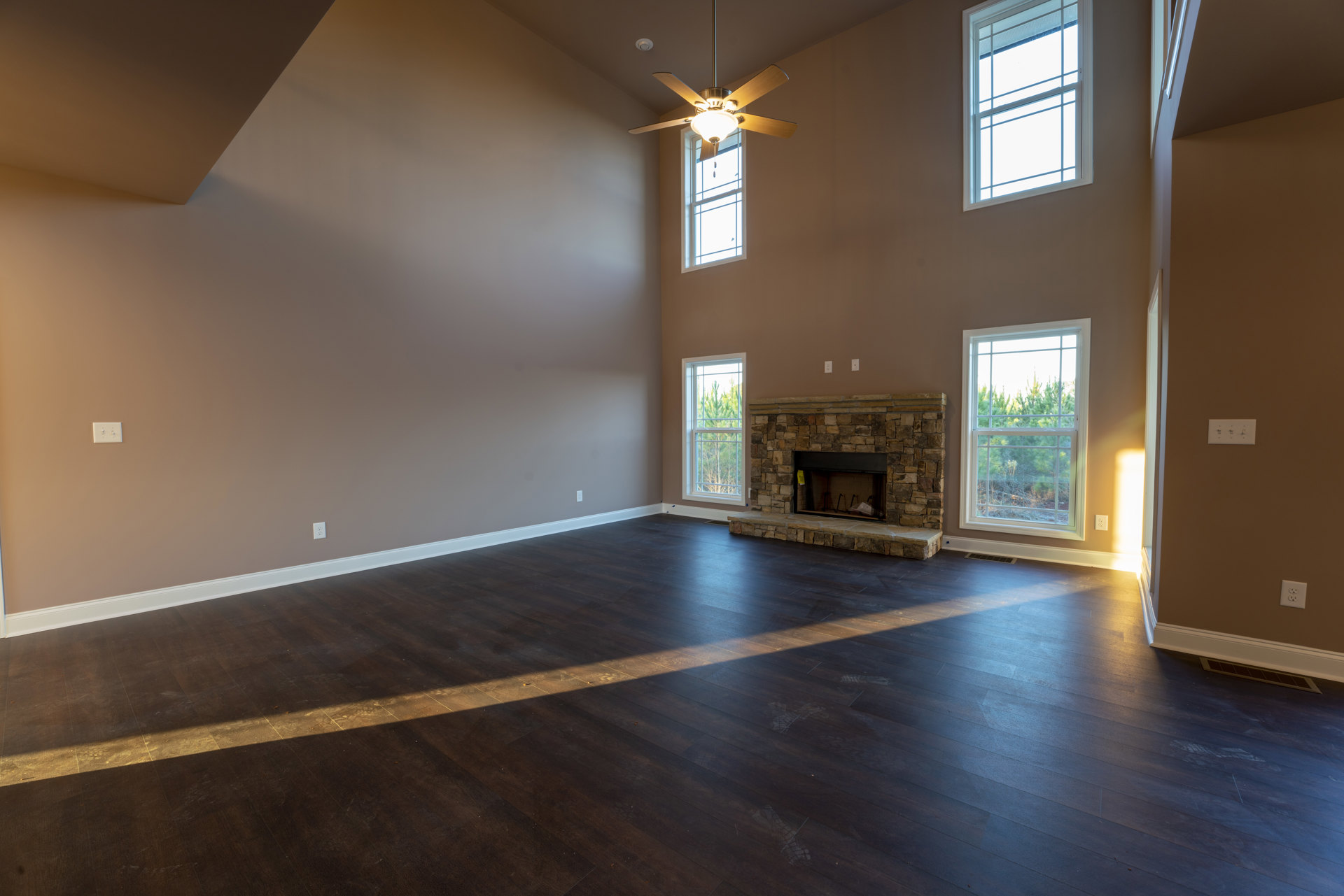 Living room with dark wood flooring, black-framed fireplace featuring a yellow tag, ceiling fan, and white-trimmed windows overlooking trees