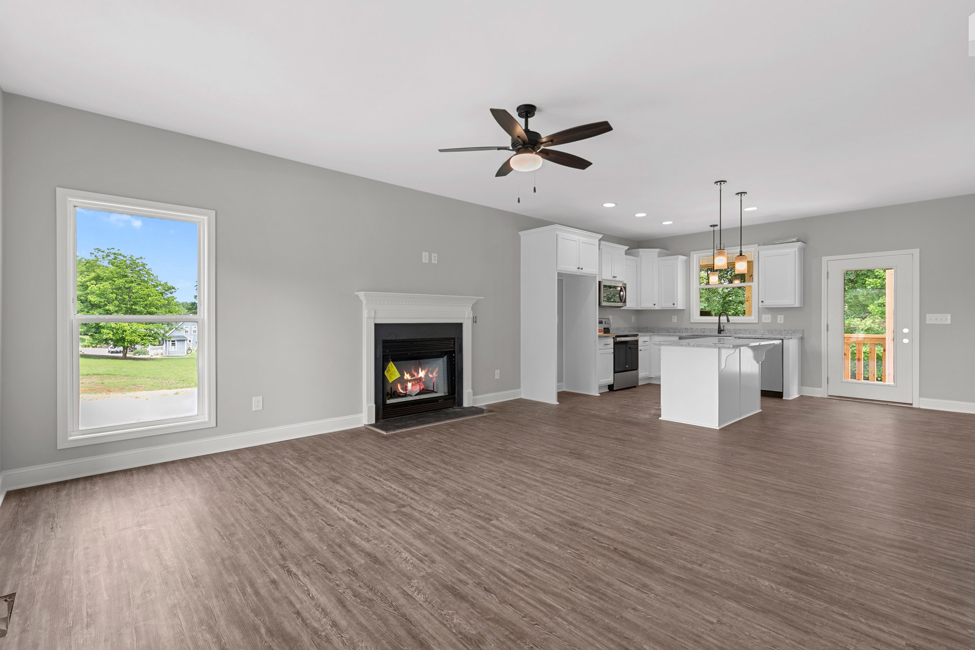 Living room with wood flooring, ceiling fan with light, stone fireplace with fire, open view into kitchen, large window showing tree and house outside