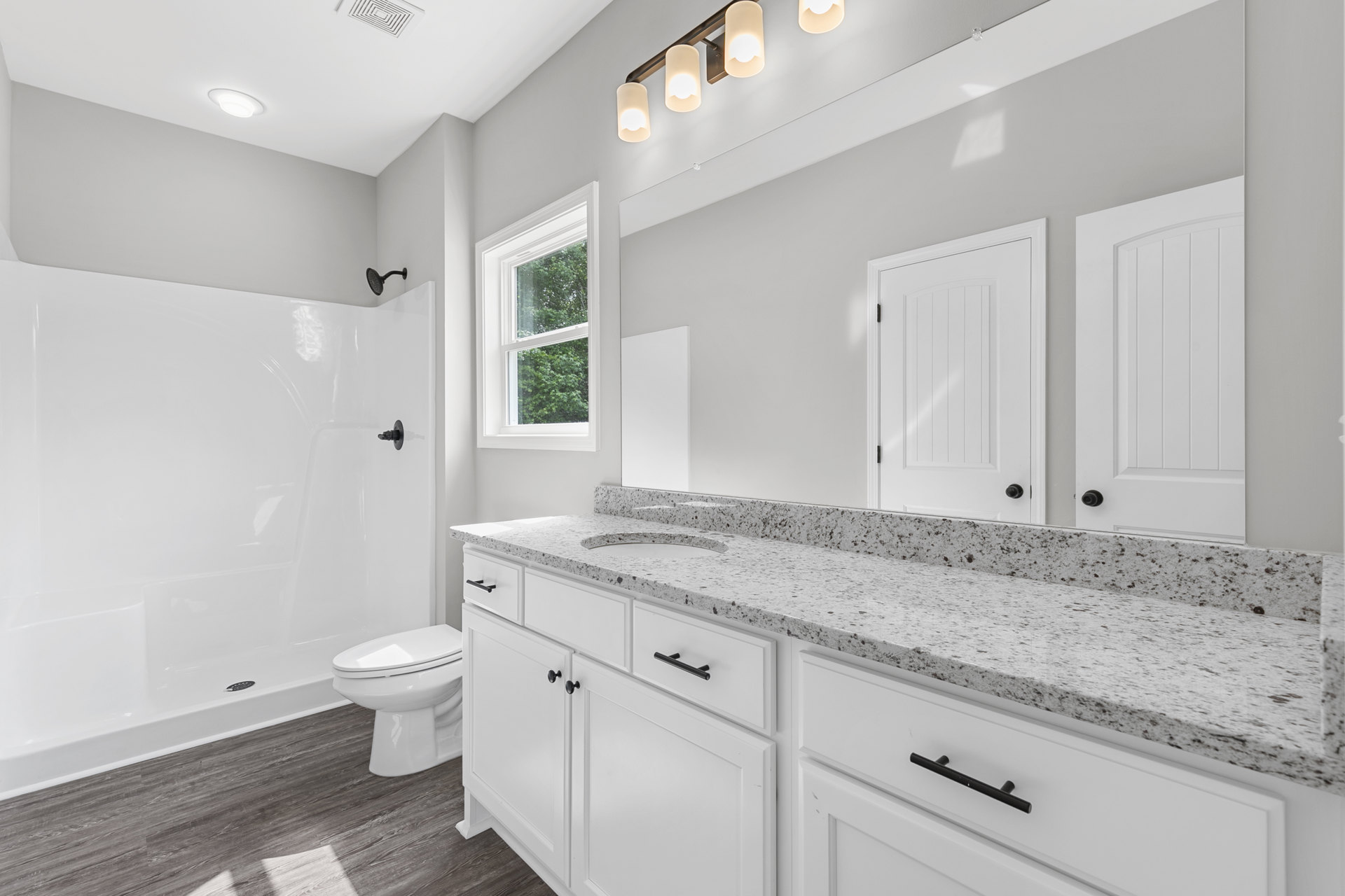 Bathroom with white shaker cabinets, white toilet, black hardware, white door with black hinges, circular ceiling light, window showing trees outside, light gray tile floor