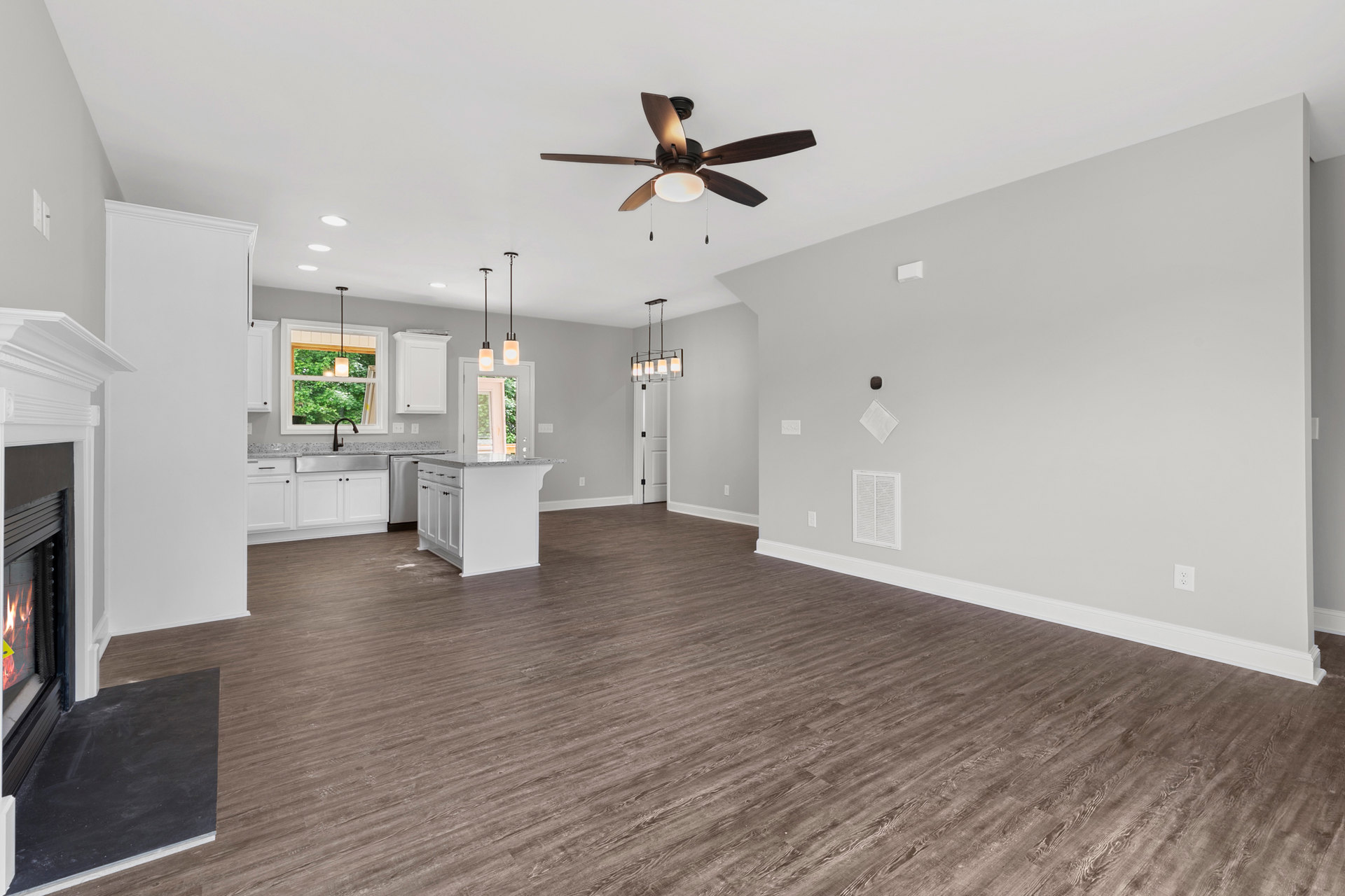 Open-concept room with wood flooring, white walls, ceiling fan with light, white kitchen island topped with marble, window letting in natural light, and white vent on ceiling.