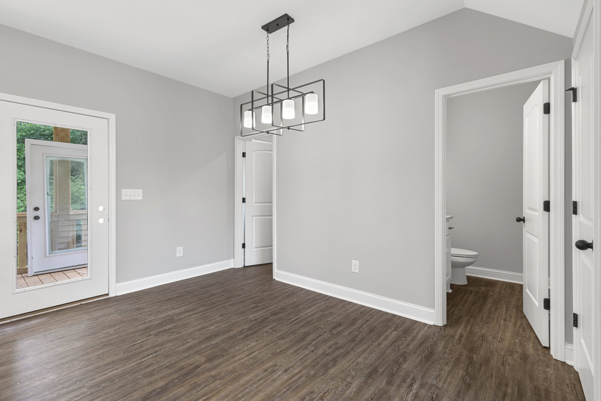 Wood flooring with white trim, ceiling-mounted light fixture, white door featuring a glass panel, window set in wood-paneled wall, partial view of a toilet in adjacent space