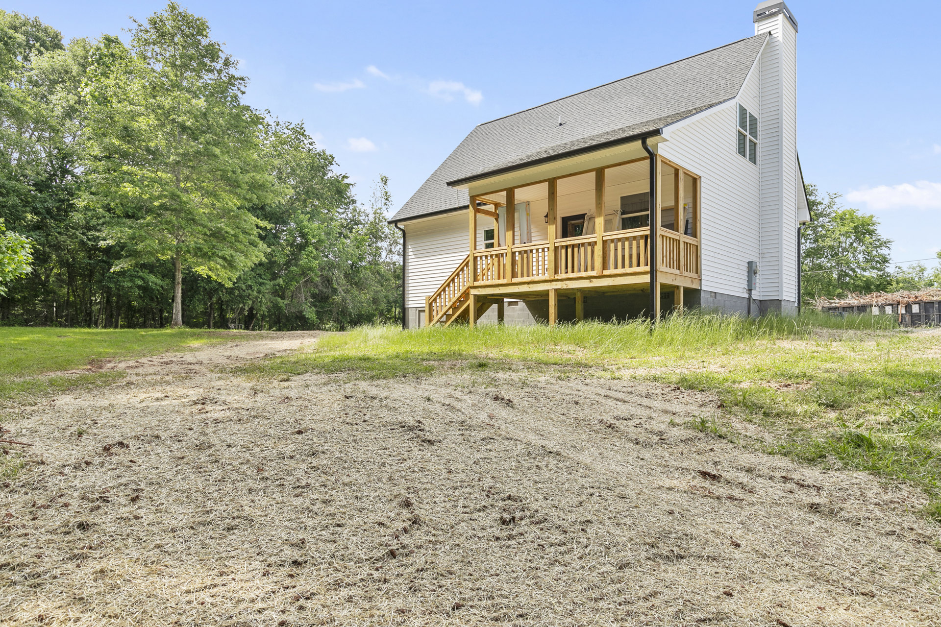 White cottage-style house with covered front porch, gravel walkway, grassy yard, and mature trees in the background
