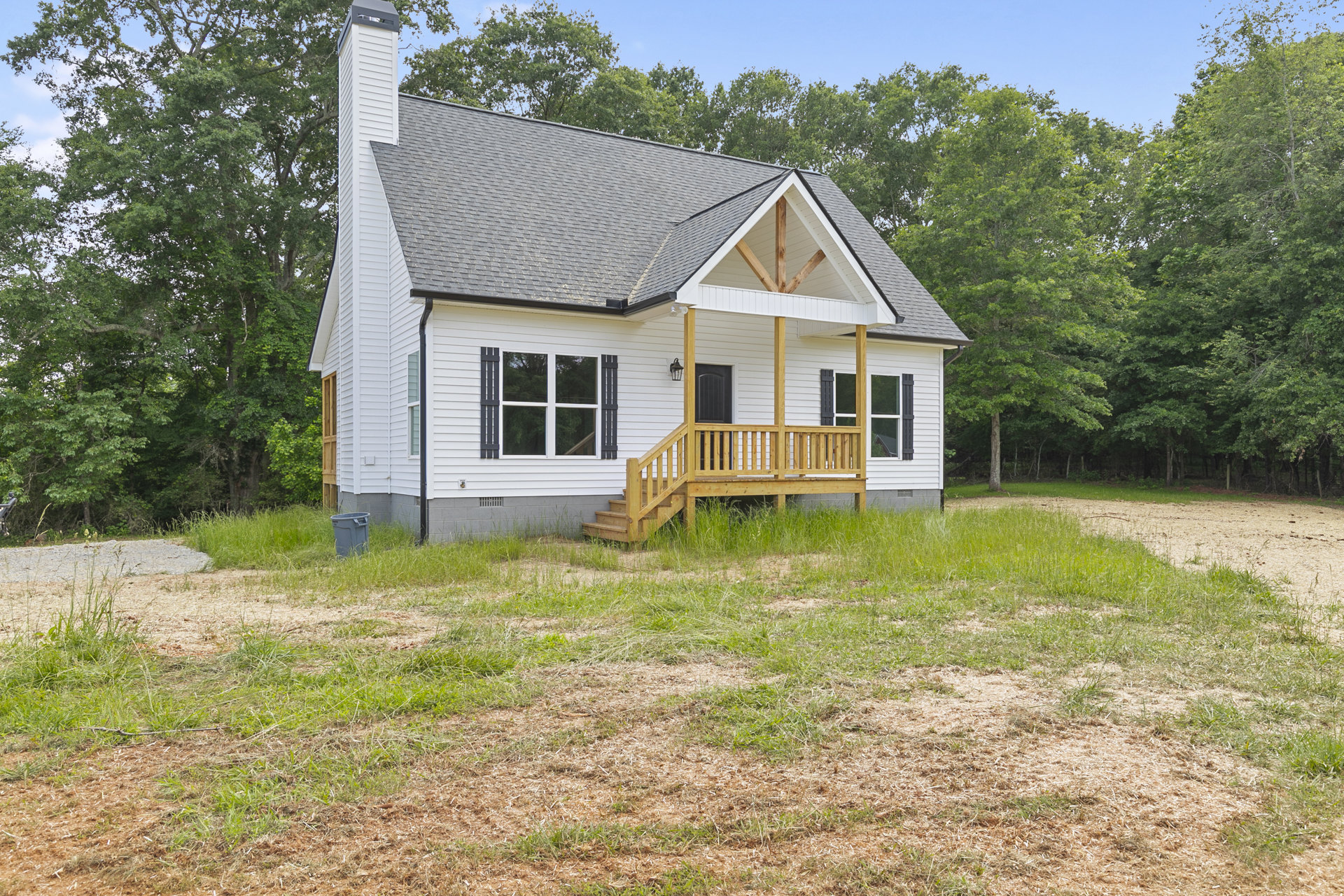 White house with a wood-railed porch, black front door, white-framed windows, wooden deck, grassy yard, grey trash can, and trees in the background