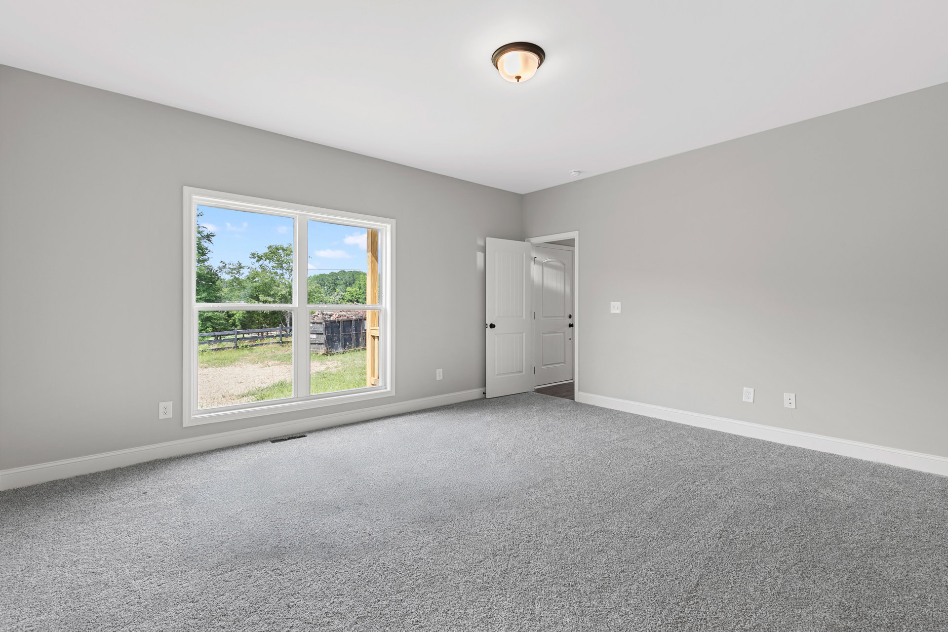 Carpeted bedroom with white walls, large window overlooking trees and fence, white door with black hardware, and ceiling light fixture