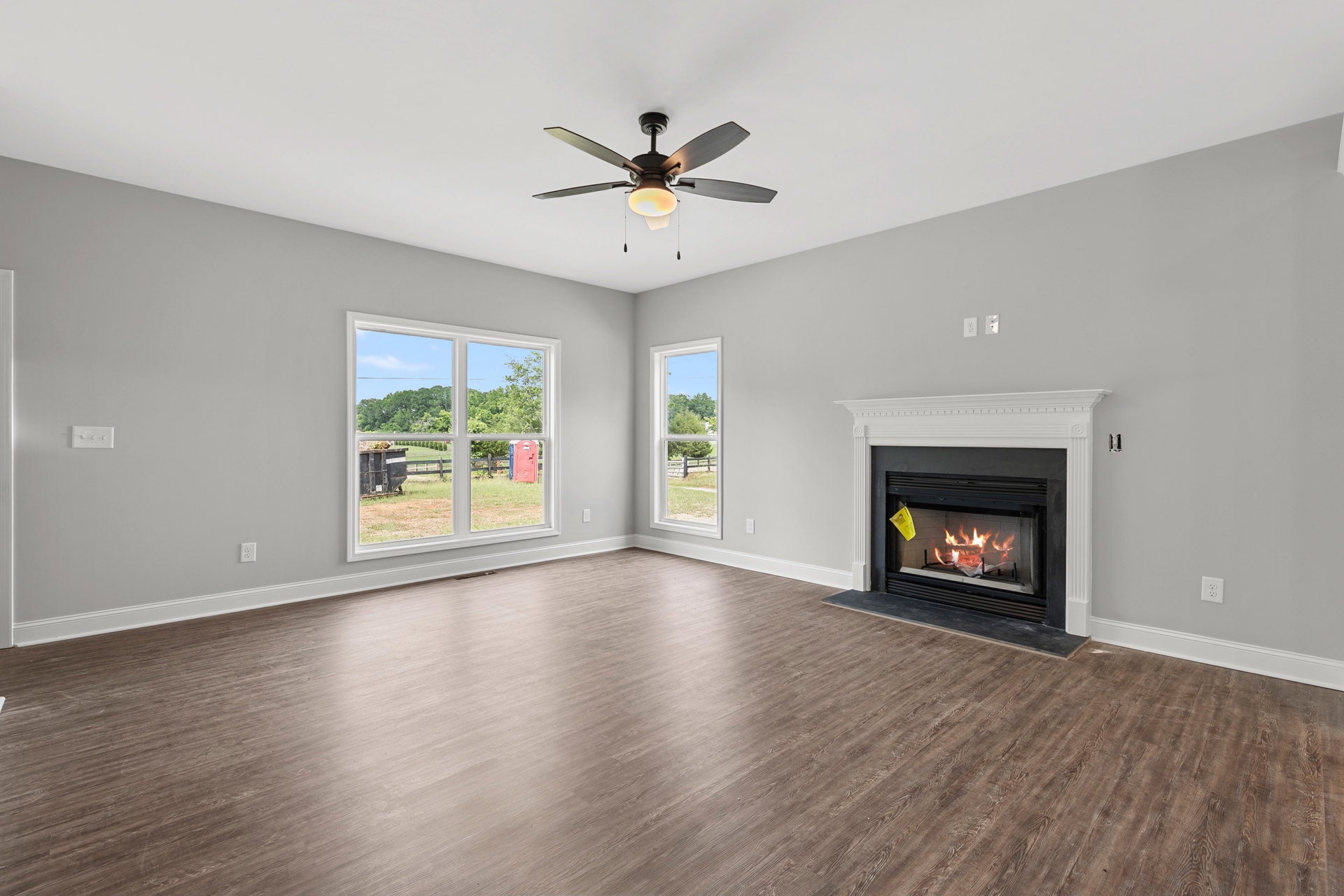 Living room with wood flooring, stone fireplace with fire, ceiling fan with light, large windows showing fenced yard and trees