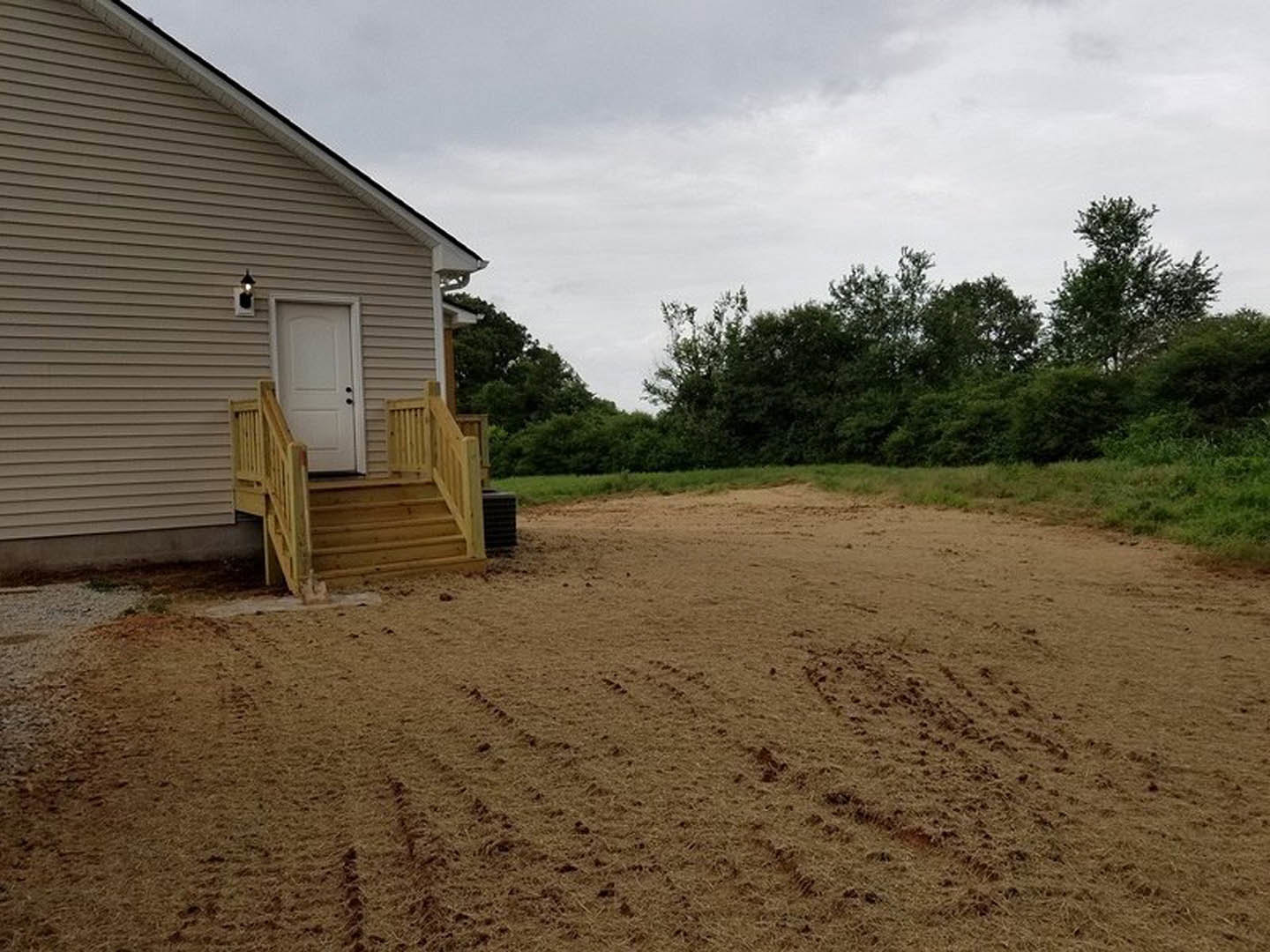 Wooden stairs leading to a cottage-style house with a white door and black knobs, surrounded by a dirt yard and scattered trees under a cloudy sky.