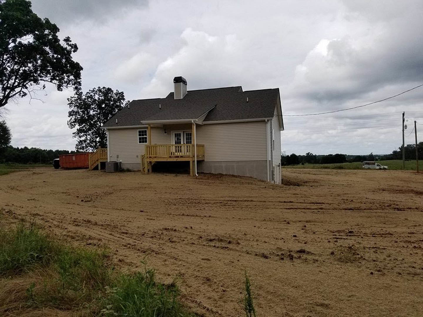 Two-story house with covered porch and wooden deck, surrounded by a large dirt field, set against a cloudy sky with sparse trees and grass.