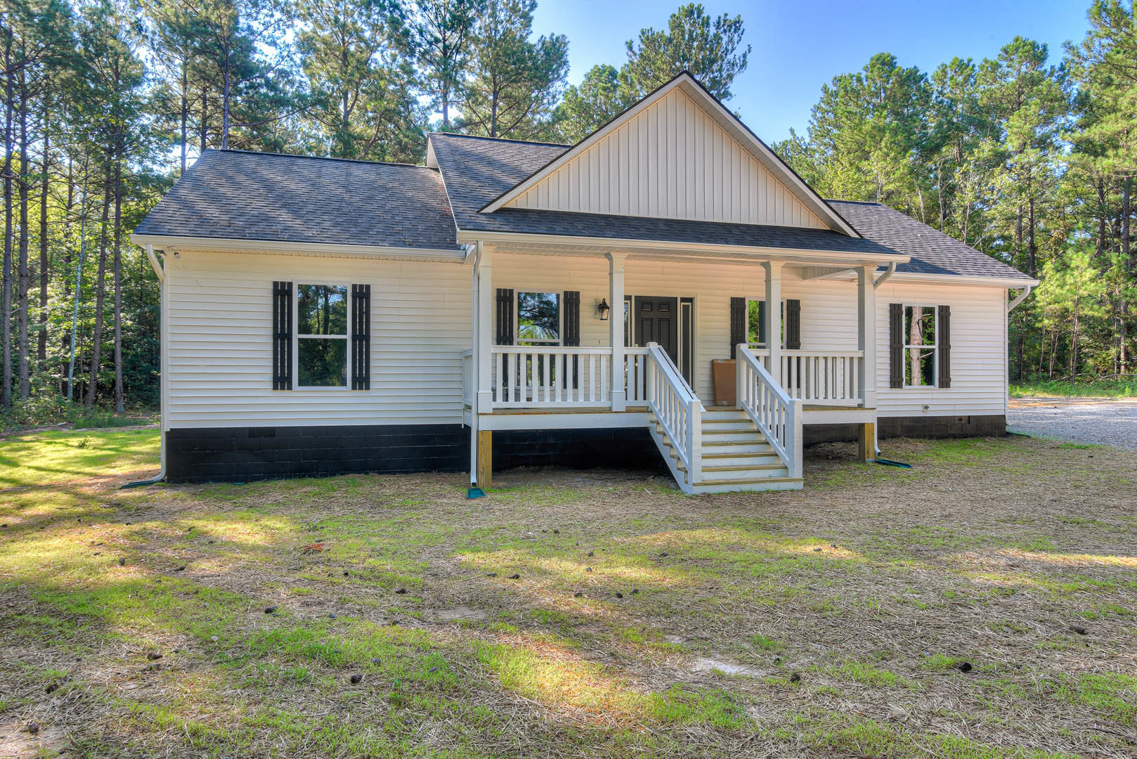White siding home with black shingle roof, covered front porch, white railings and stairs, black shuttered windows, grassy lawn, and mature tree nearby