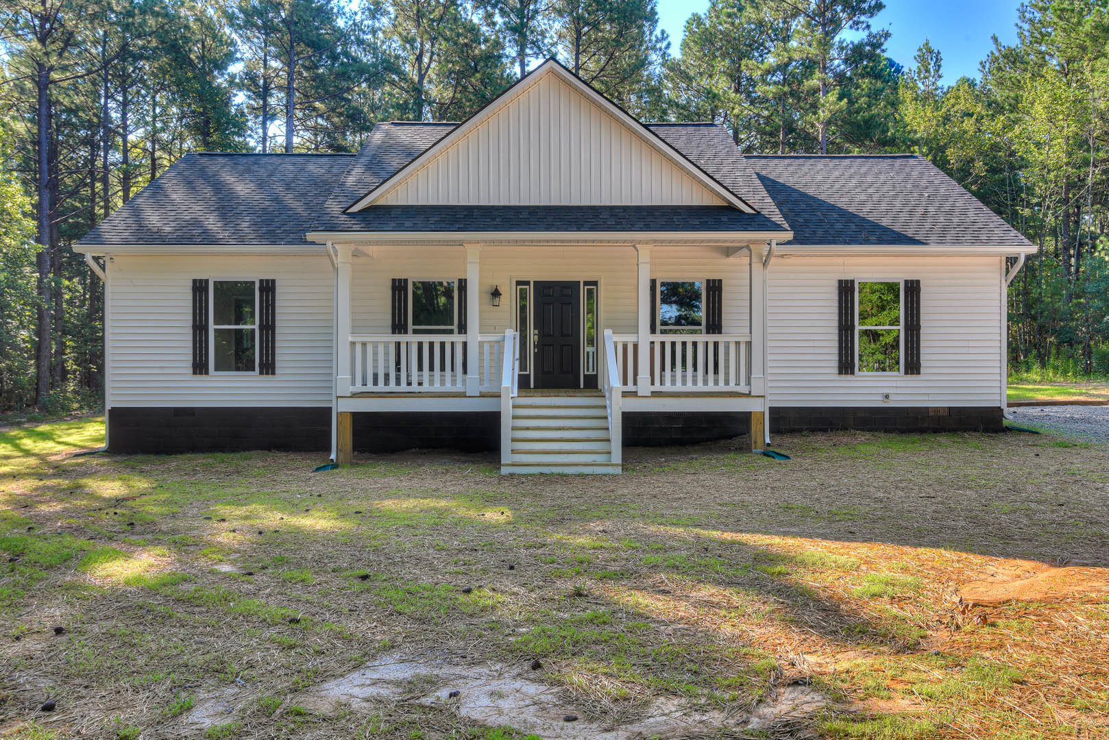 White cottage-style home with black door and glass panels, black window shutters, grassy front yard, mature trees in background.