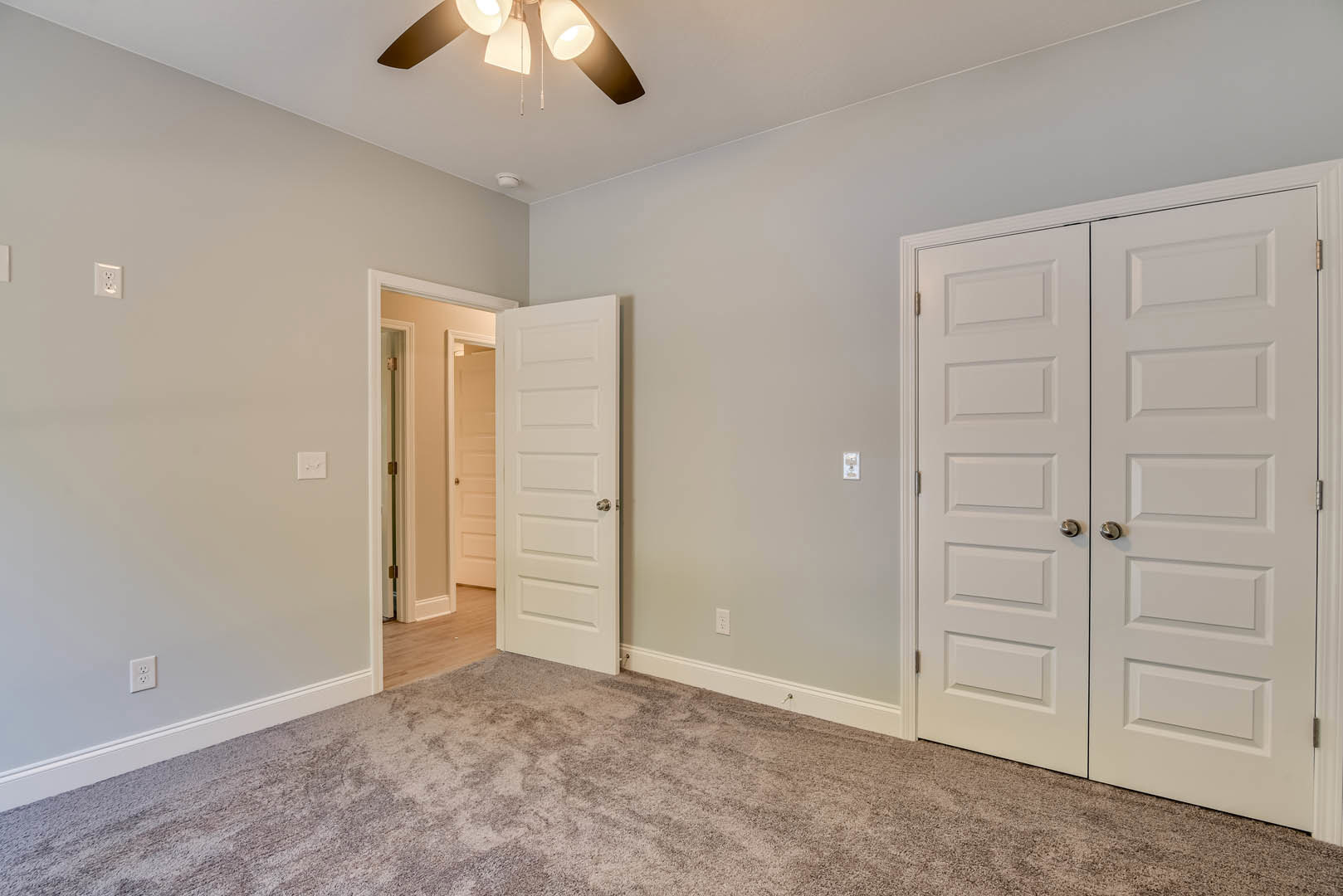 Bedroom with light carpet flooring, white double doors featuring silver knobs, and a ceiling fan mounted on a white plaster ceiling.