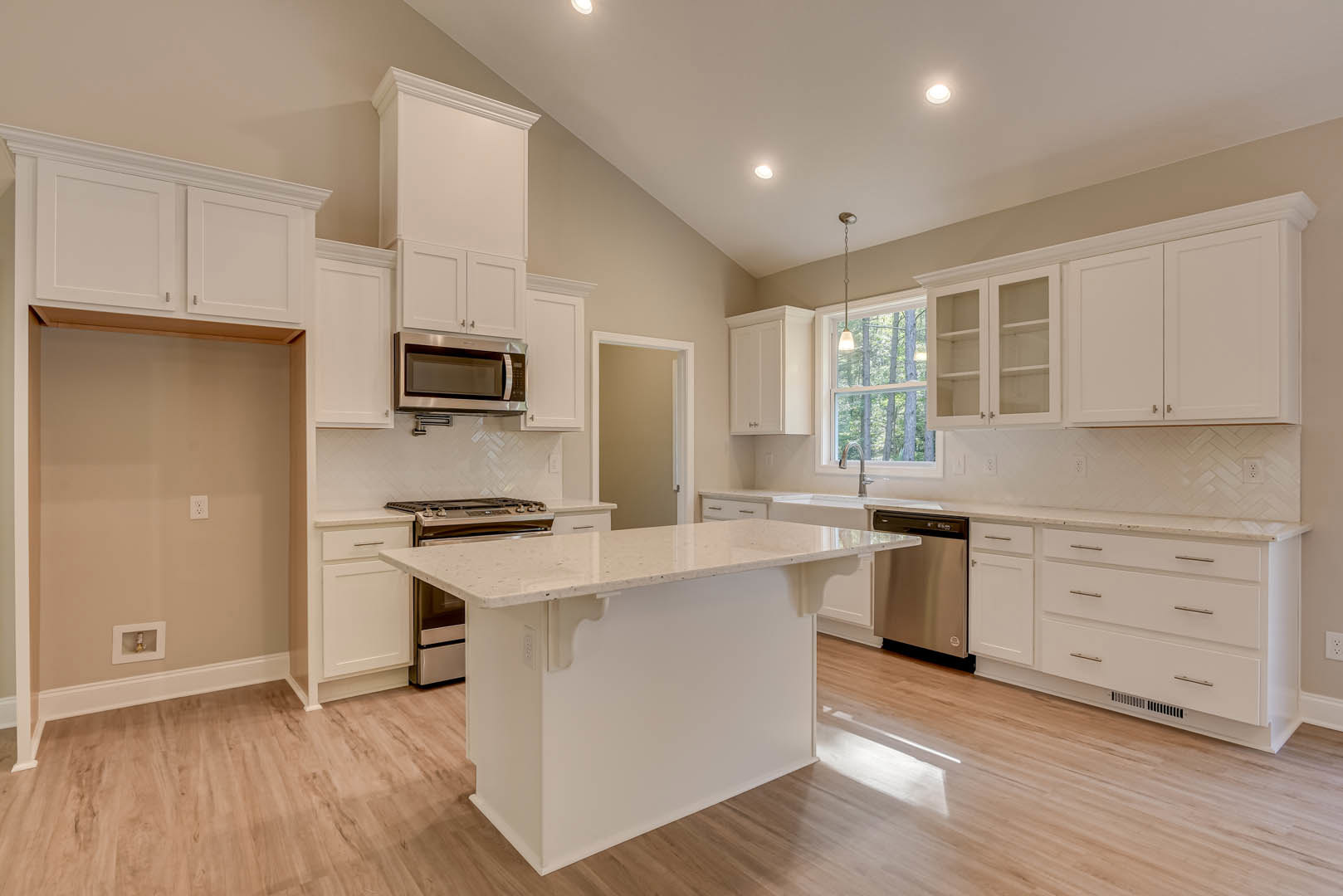 White kitchen with shaker cabinets, large central island with quartz countertop, stainless steel appliances, wood flooring, open shelving, and a wall-mounted light switch.