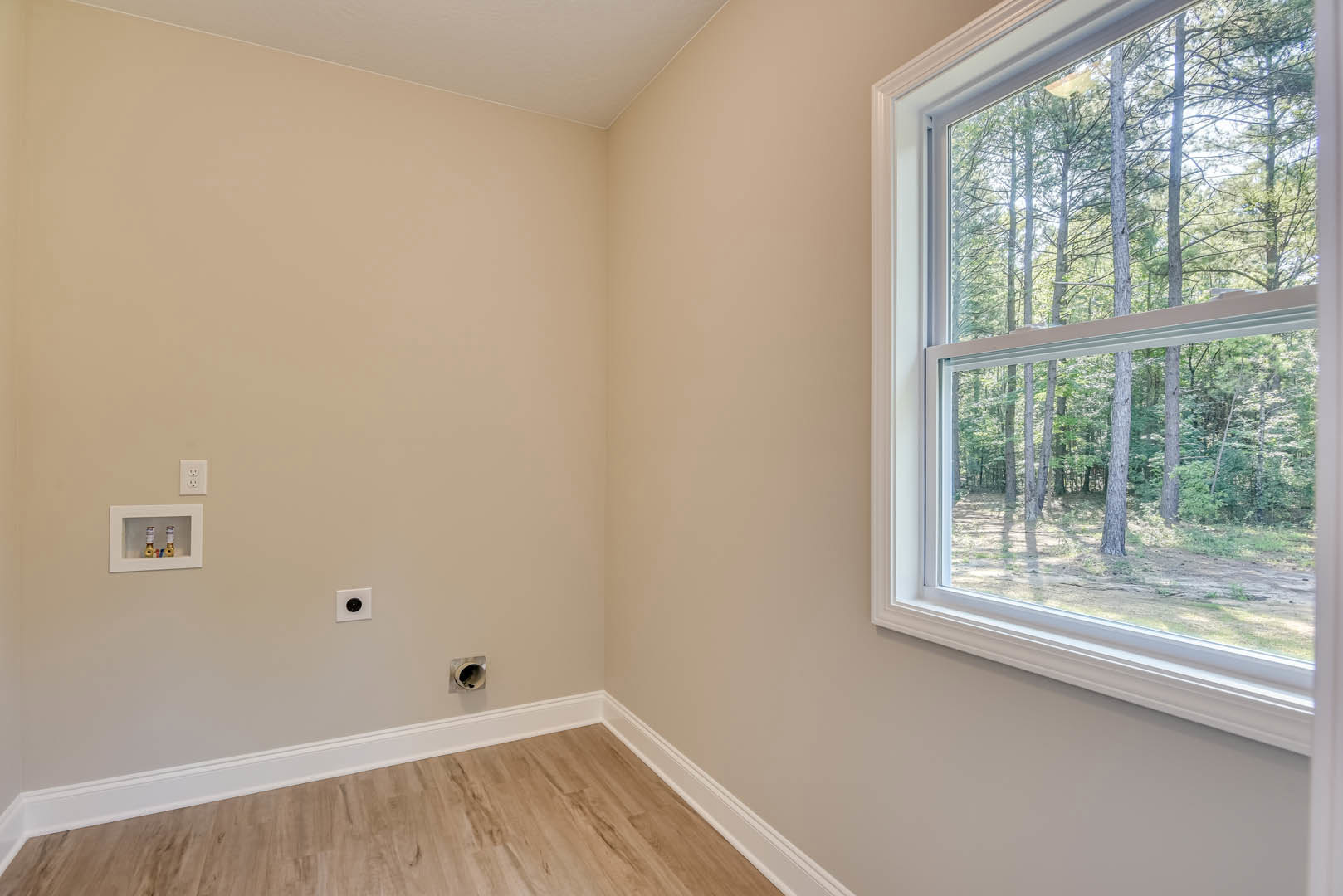 Sunlit room featuring a large window with views of green trees, smooth white walls, wood laminate flooring with white baseboards, recessed ceiling detail, and a white cabinet