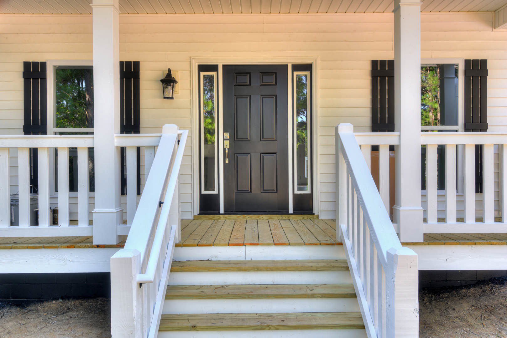 Black front door with glass panels, white railing and balusters, black outdoor lamp, window reflecting trees, gray porch steps