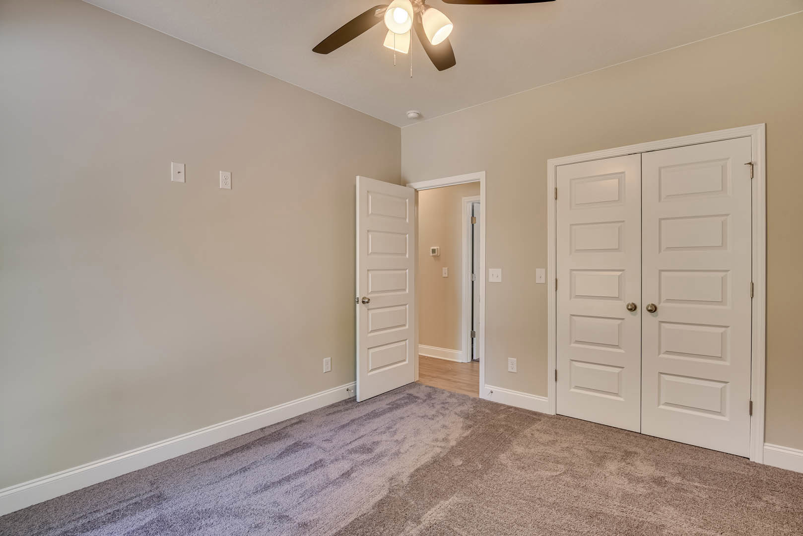 White double doors with silver knobs, beige carpeted floor with visible stain, ceiling fan and light fixture, smooth white walls and ceiling.