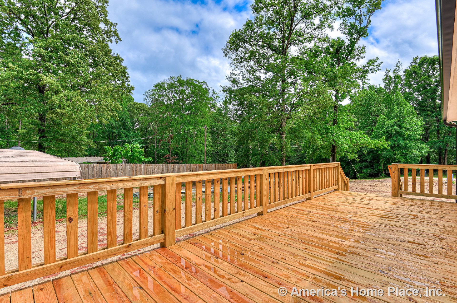 Expansive wooden deck with natural finish, featuring vertical slat railings and open layout, overlooking a fenced backyard with mature trees.